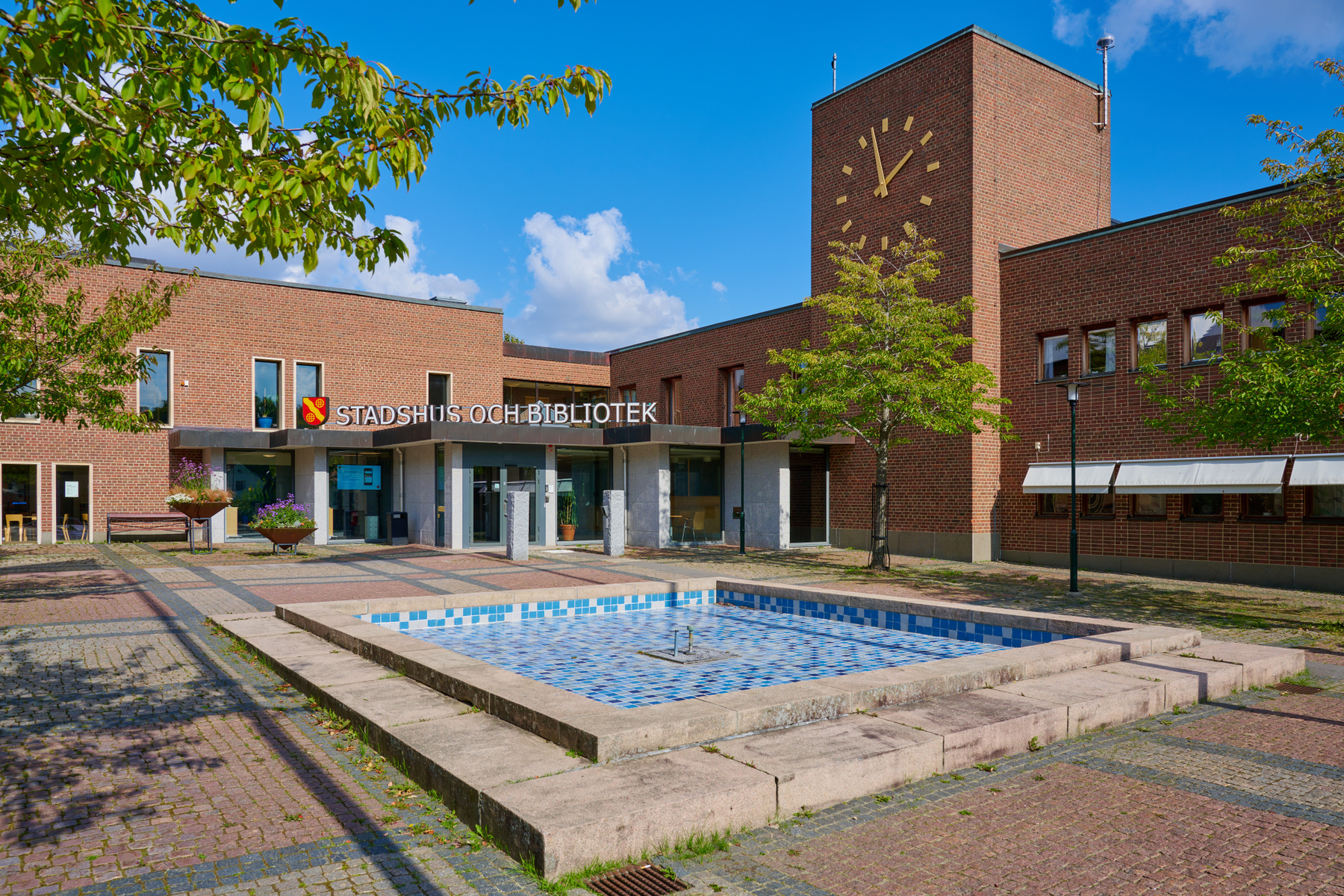 En fasadvy av en tegelbyggnad med texten "STADSHUS OCH BIBLIOTEK" ovanför ingången. Till vänster i bilden syns trädgrenar med gröna löv och i förgrunden finns en kvadratisk fontän med blått och vitt mosaikmönster. Byggnaden har ett klocktorn med guld