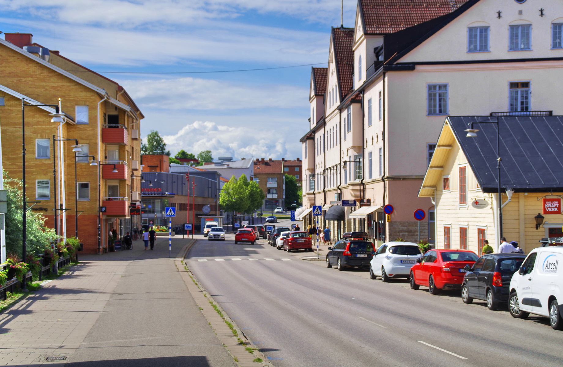 En gata i en stad på en solig dag, kantad av byggnader i olika färger som gult och rosa. Flera bilar är parkerade längs gatan, och en del människor går på trottoarerna. Blå himmel med vita moln syns ovanför byggnaderna.