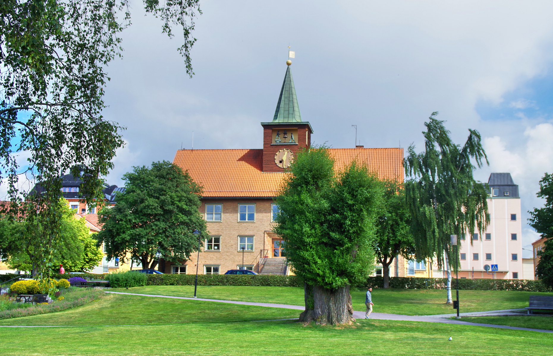 En bild som visar en gräsplätt i förgrunden, med träd och en byggnad bakom. Byggnaden har ett tegeltak och ett grönfärgat torn med klocka. På gräsplätten syns en person på en gångväg intill ett träd.