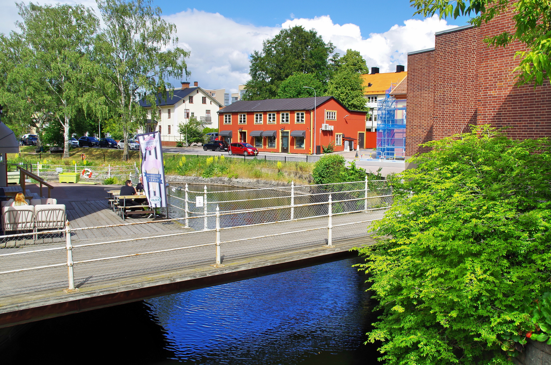 En översiktsbild av en bro över en kanal med byggnader, träd och människor. En röd byggnad och en tegelbyggnad syns i bakgrunden. Ett par sitter vid ett bord på en uteservering vid kanalbrädden.