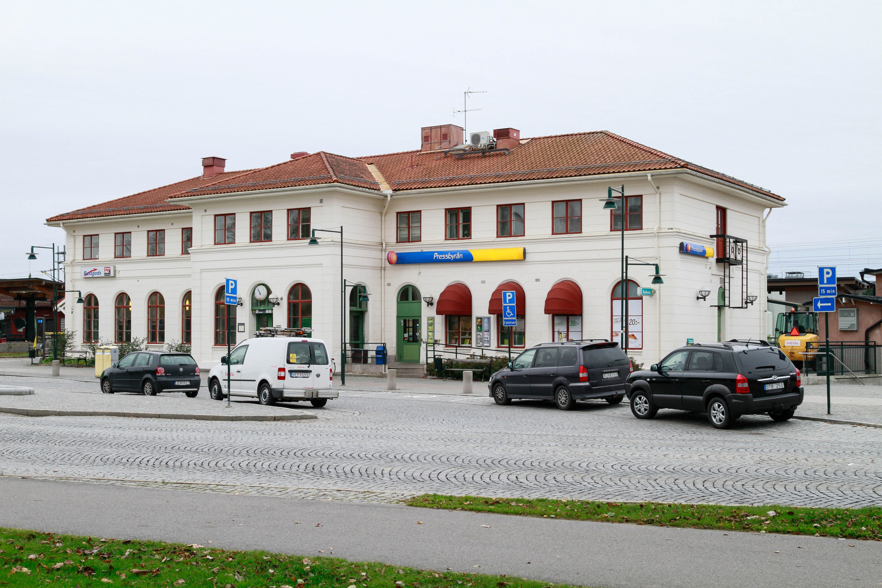 En bild som visar en ljus byggnad med rött tak framifrån, med bilar parkerade på kullerstensbelagd mark framför. Skyltar med texten "Pressbyrån" och parkeringsskyltar syns tydligt. En grön gräsremsa med vissna löv kan ses i förgrunden.