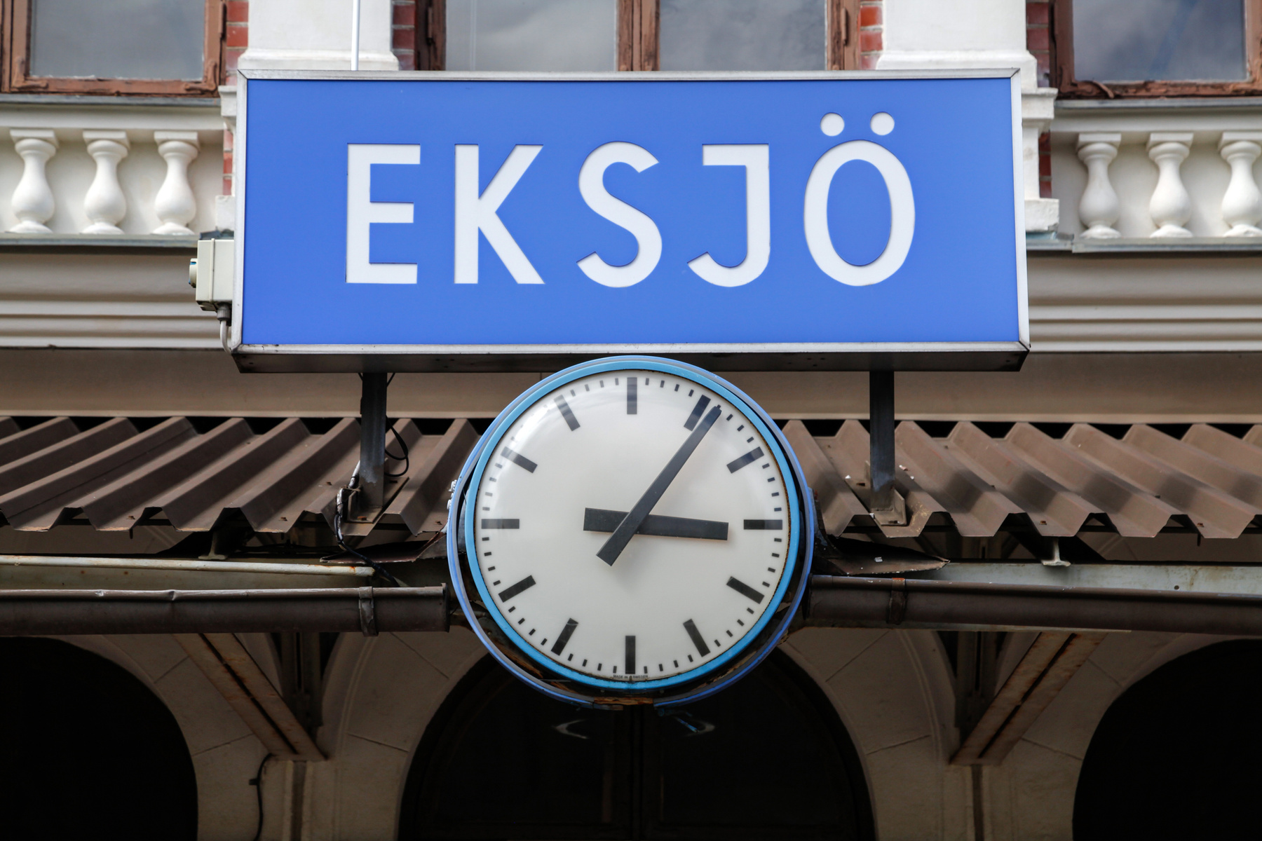 En blå skylt med texten "EKSJÖ" i vitt hänger ovanför en rund, vit stationsklocka med svarta visare och markeringar. Bakom skylten och klockan syns en del av en byggnad med fönster och en takkonstruktion, samt en mörkblå himmel.