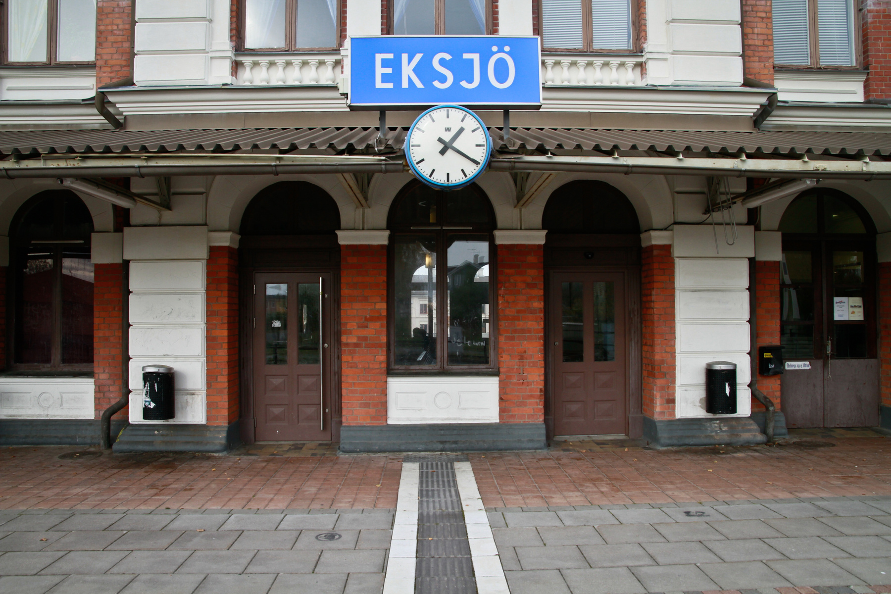 En fasad på en byggnad med tegel och vita detaljer. Ovanför entrén hänger en blå skylt med texten "EKSJÖ" och under den sitter en rund klocka. Tre bruna dörrar och två fönster med valvformade överdelar syns på fasaden.