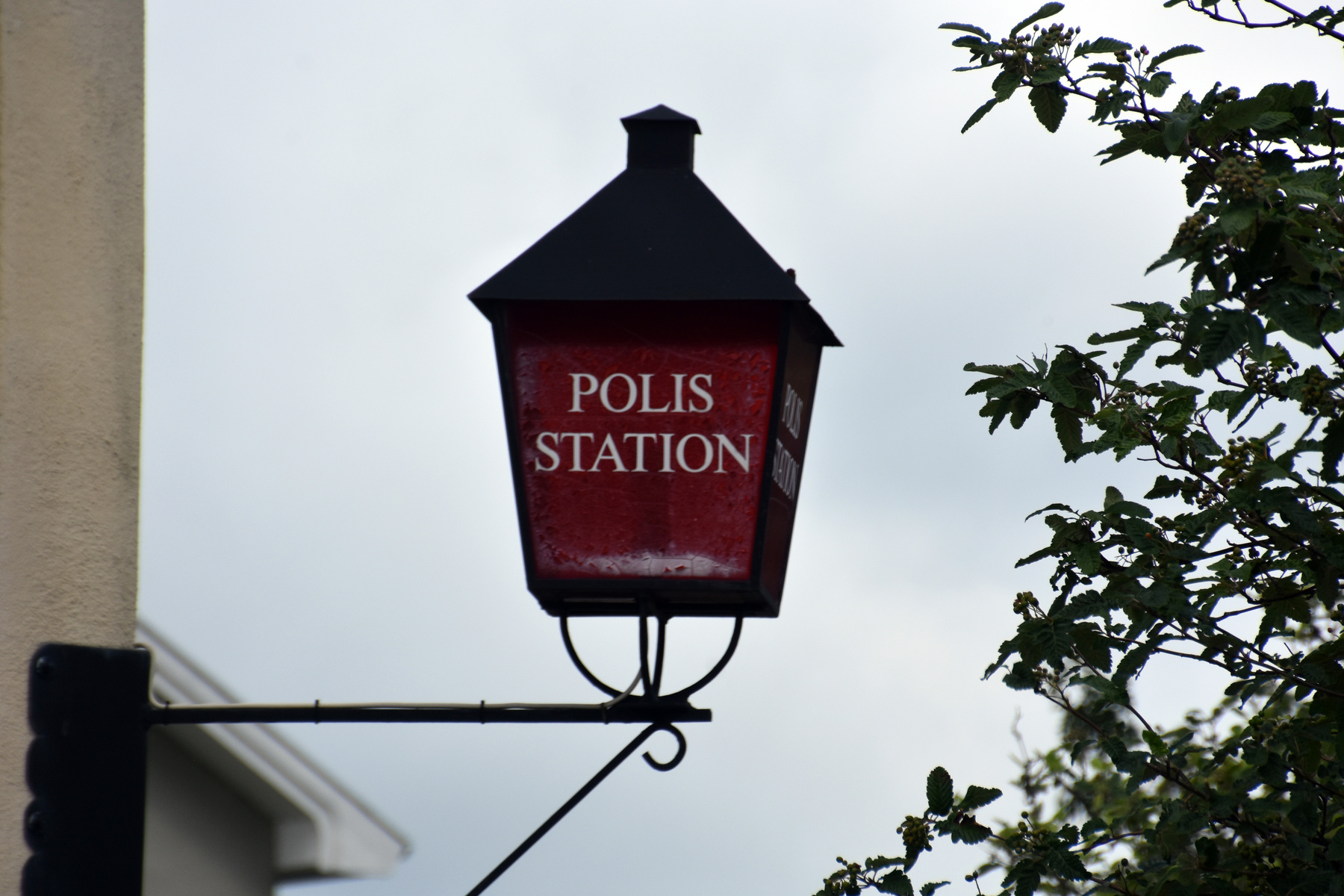 En röd och svart skylt med texten \"POLIS STATION\" hänger fäst vid en byggnad mot en grå himmel. Till höger om skylten syns gröna löv och grenar från ett träd. Skylten är fäst i en svart metallarm.