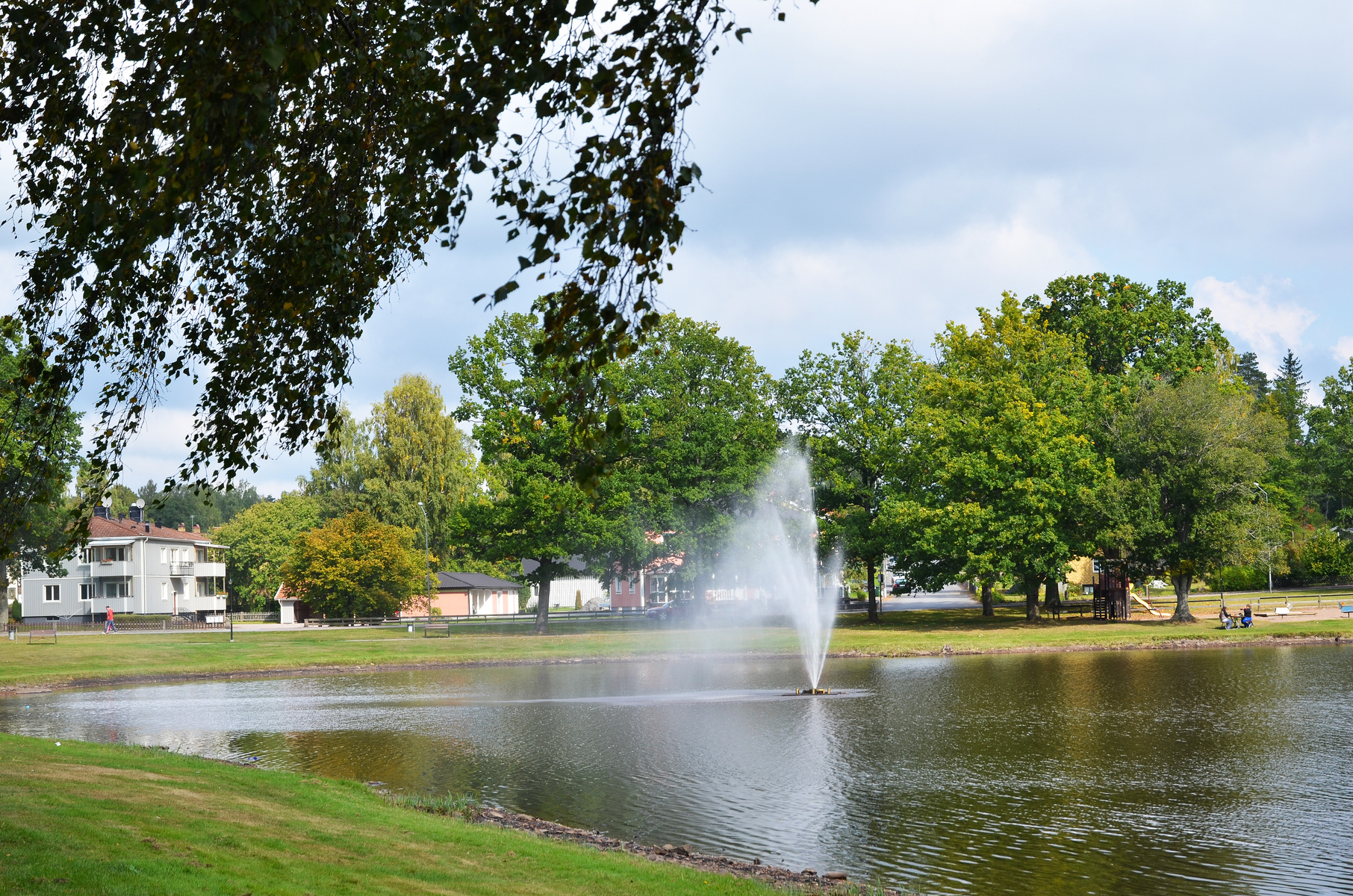 En bild som visar en sjö med en aktiv fontän i mitten. Runt sjön finns gräsytor och träd. I bakgrunden syns bostadshus och andra byggnader under en delvis molnig himmel. Två personer sitter bland träd på höger sida av sjön.