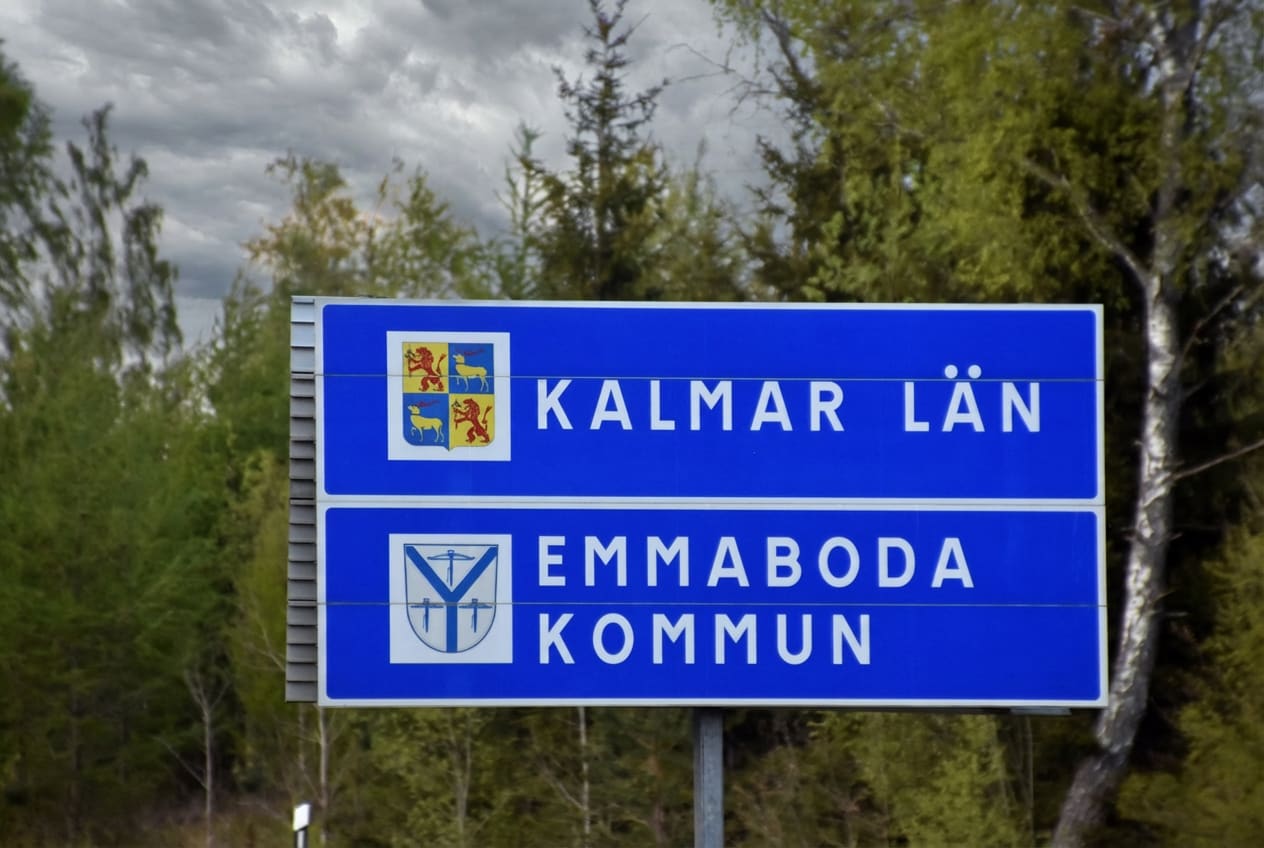 En blå vägskylt med vit text som visar "KALMAR LÄN" och "EMMABODA KOMMUN". Skylten har även respektive kommuns vapensköldar. Bakgrunden består av gröna träd och en gråmulen himmel.