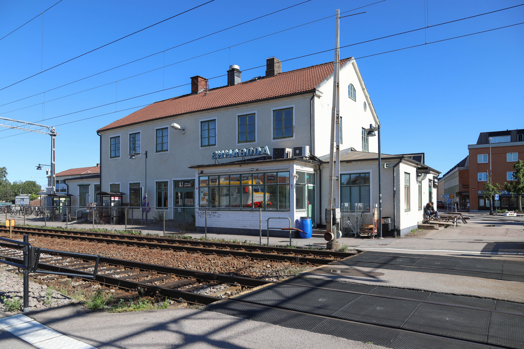 En dagbild med ett stationshus med texten "EMMABODA" på taket. Framför huset, över spåren, syns en järnvägsövergång. Huset har rött tak och ljusa fasader samt gröna fönsterramar. Två personer syns sitta på en träbänk till höger om stationshuset.