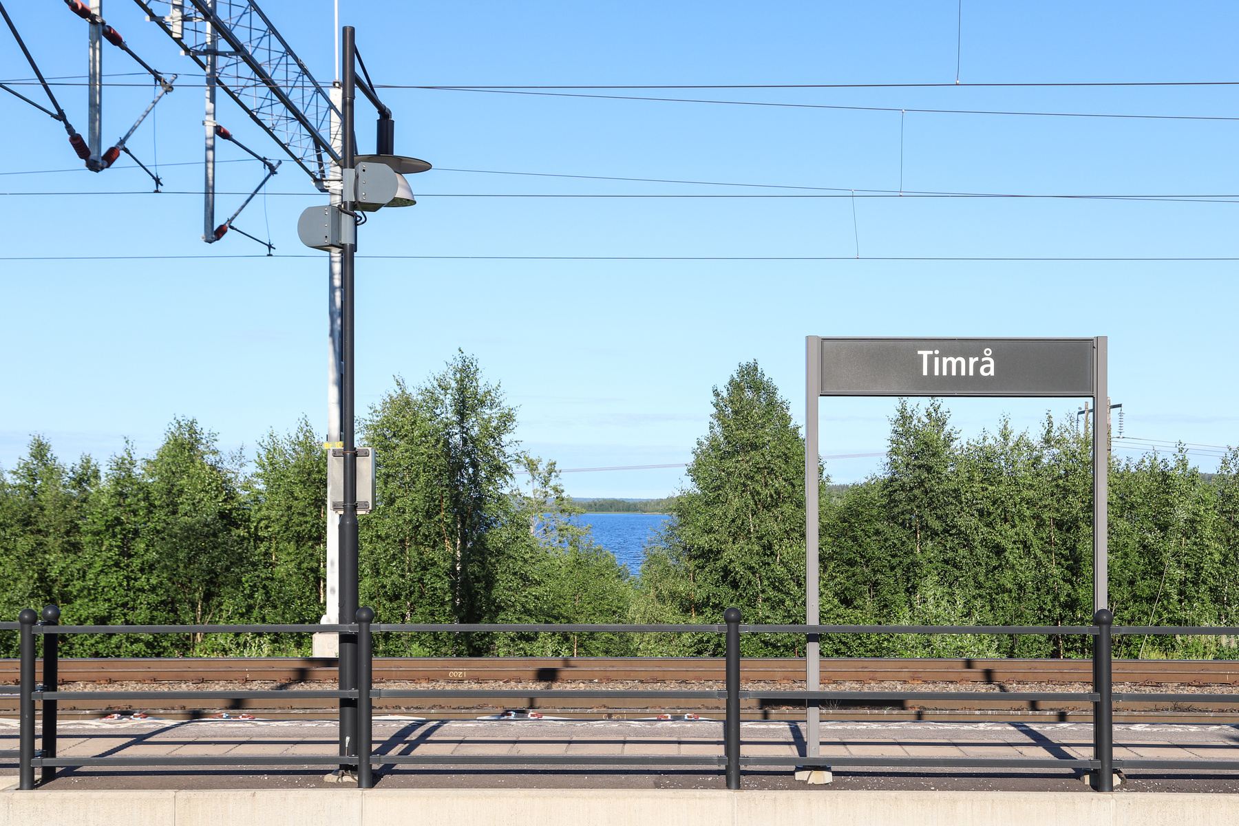 En bild av en tågplattform med en skylt som säger "Timrå". Bakom plattformen syns tågspår, grönskande träd och ett vattendrag under en klarblå himmel. Över spåren finns ett nät av elledningar och en kontaktledningsstolpe.
