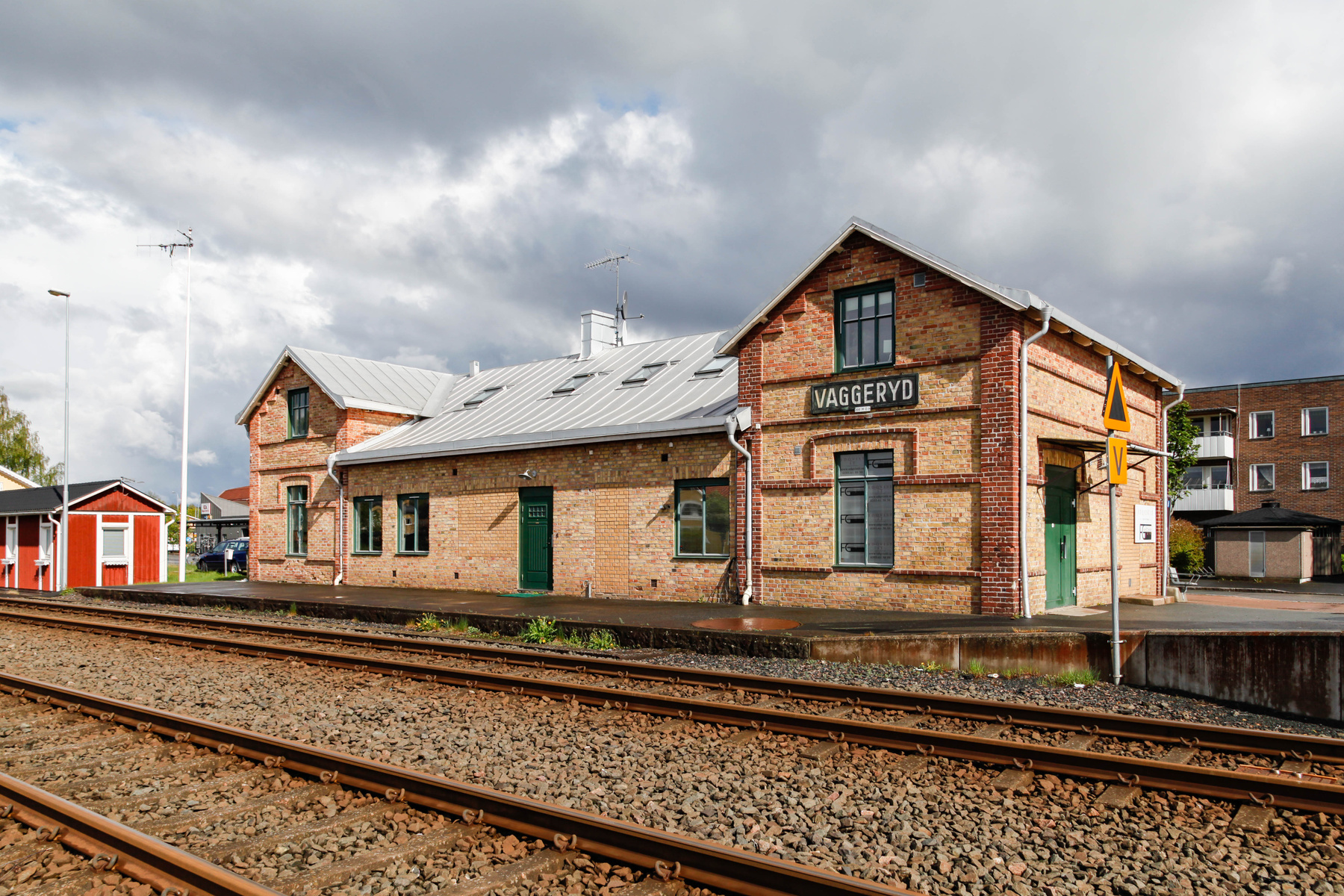 En gulbrun tegelsbyggnad med ordet "VAGGERYD" skrivet i versaler på fasaden står bredvid två järnvägsspår. Byggnaden har fönster och dörrar med gröna detaljer samt ett grått plåttak. I bakgrunden syns molnig himmel, ytterligare en röd byggnad, ett fl