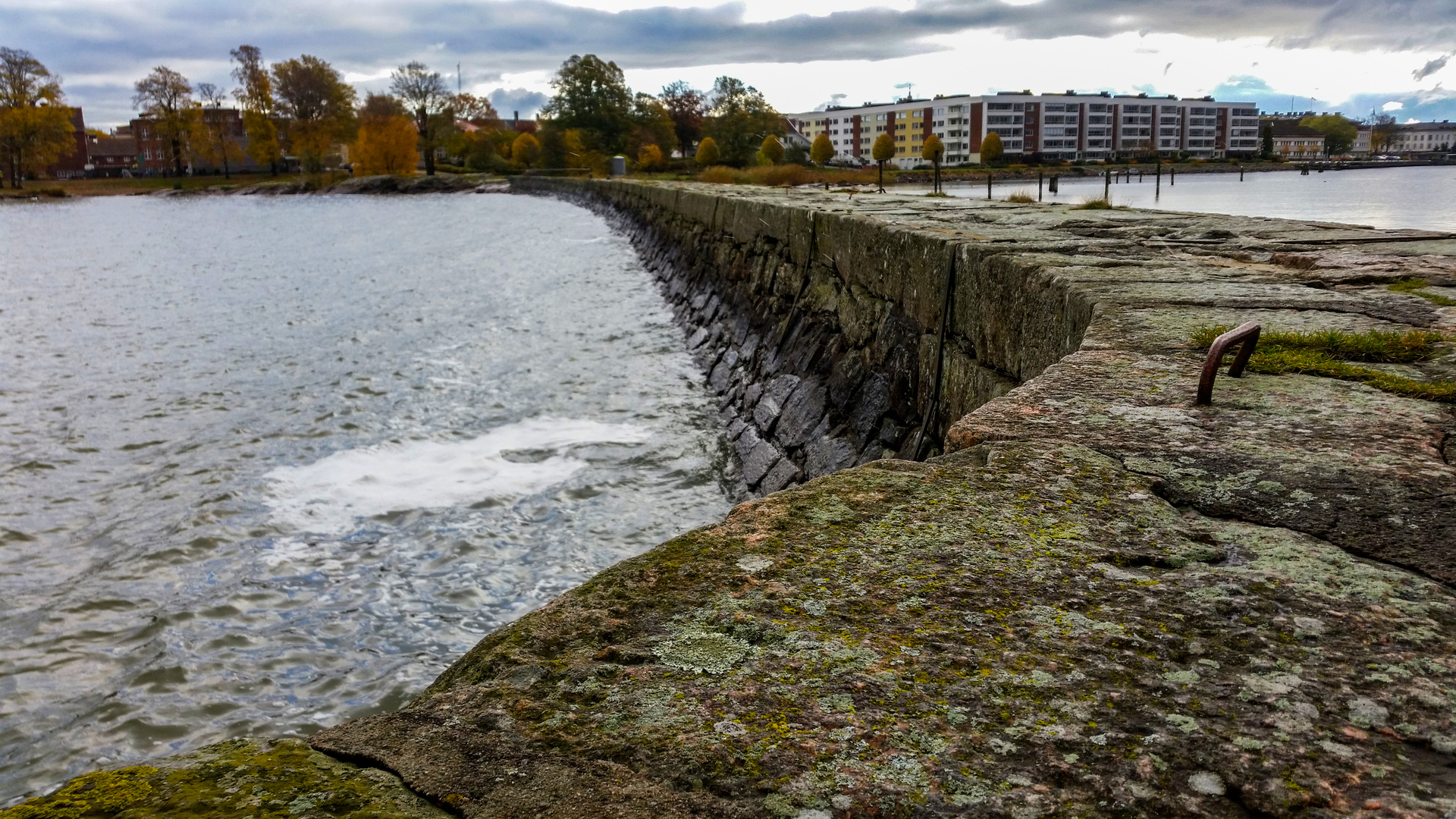 En bild som visar en stenmur som sträcker sig längs vattnet under en molnig himmel. På andra sidan vattnet syns träd med höstfärger och i bakgrunden skymtar byggnader. Framträdande i förgrunden är mossbevuxen sten och en rostig metallögla.