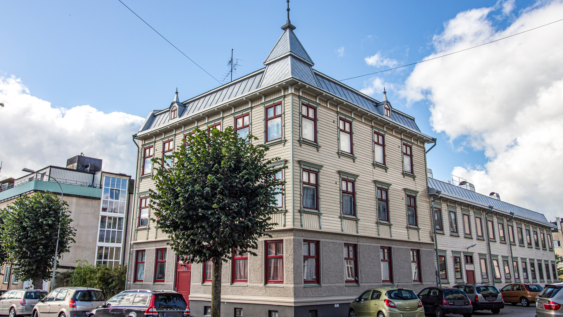 En stor, ljusbeige byggnad med mörkröda fönsterkarmar och en grå takkonstruktion med en spetsig del i mitten syns under en ljusblå himmel med vita moln. Framför byggnaden står parkerade bilar och ett träd med gröna löv.