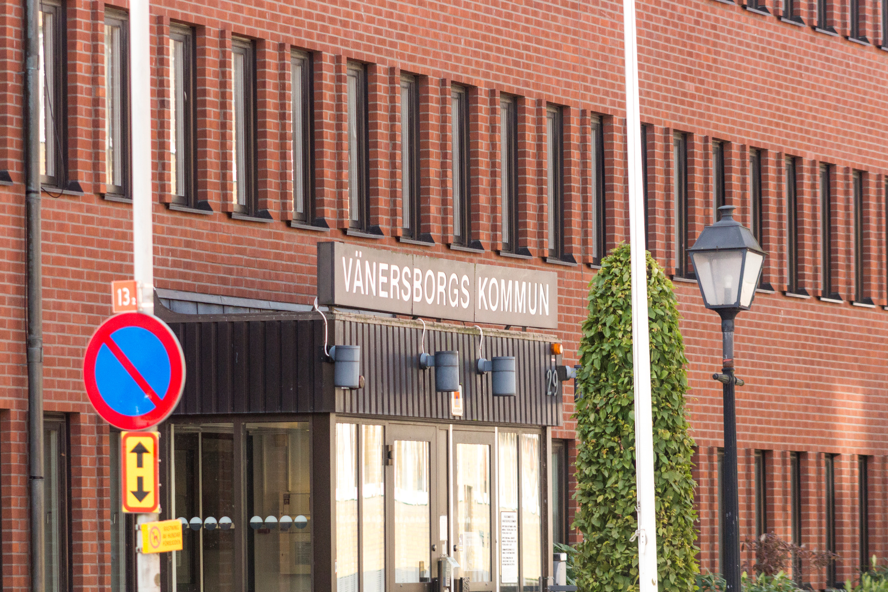 En bild av utsidan på ett tegelhus med texten "VÄNERSBORGS KOMMUN" ovanför ingången. Framför byggnaden syns trafikskyltar, en lyktstolpe, en flaggstång och en grön buske.