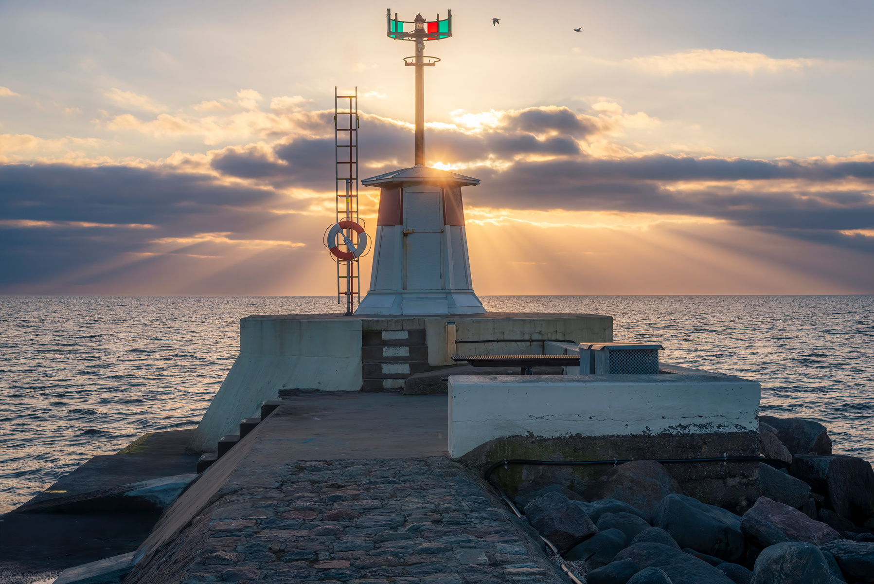 En fyrbåk på en brygga vid havet i solnedgången. Solstrålar sprider sig genom molnen. Vattenlinjen sträcker sig horisontellt, medan fåglar flyger i skyn. En livboj och en stege syns bredvid fyren.