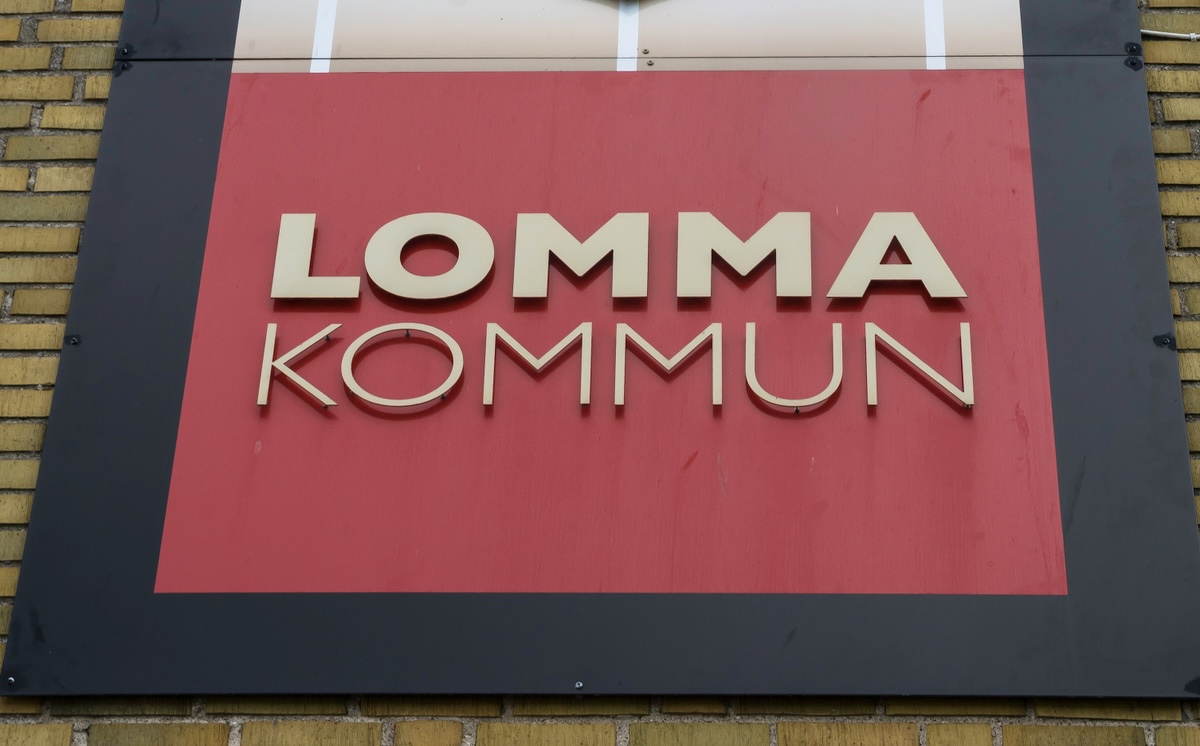 En skylt för "LOMMA KOMMUN" med guldfärgade bokstäver på en röd bakgrund, monterad på en byggnad med gult tegel. Skylten har en svart ram runt den röda ytan. Bilder är tagen snett nedifrån, med tegelväggen i bakgrunden till vänster och nedtill.