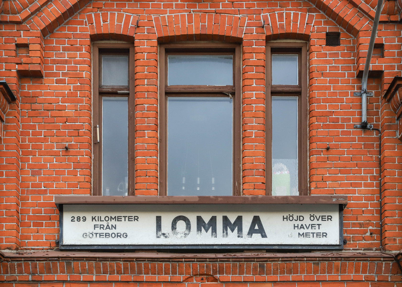 En fasad av ett hus, sannolikt en stationsbyggnad, byggd av rött tegel med tre fönster med mörkbruna karmar och en skylt under fönstren.