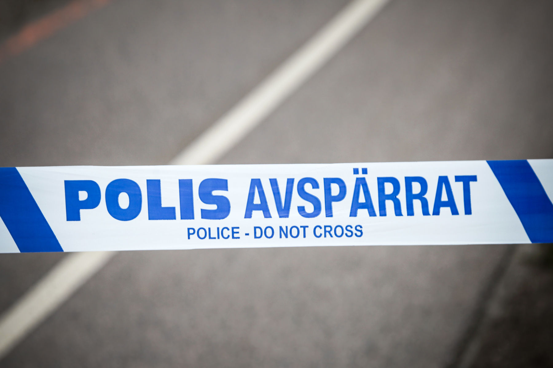 En vit och blå avspärrningstejp med texten "POLIS AVSPÄRRAT" och "POLICE - DO NOT CROSS" diagonalt över bilden. Bakgrunden är oskarp och visar en grå asfaltsyta med vita linjer som kan vara en väg eller liknande.