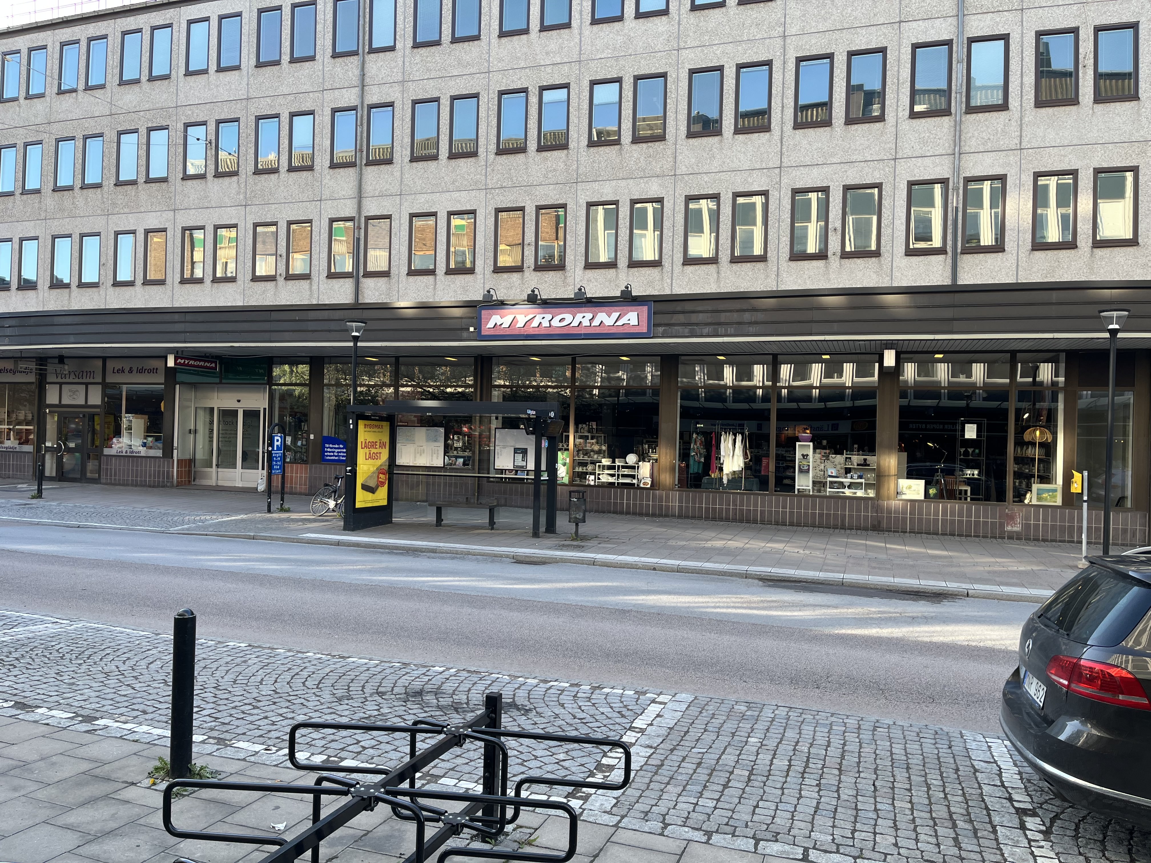 En gata med gatsten och asfalt i förgrunden, parkeringsplatser, en busskur, och kommersiella byggnader i bakgrunden. Butiker som "Värsam", "Lek & Idrott" samt "Myrorna" är synliga. Ett svart fordon skymtar i högra kanten.