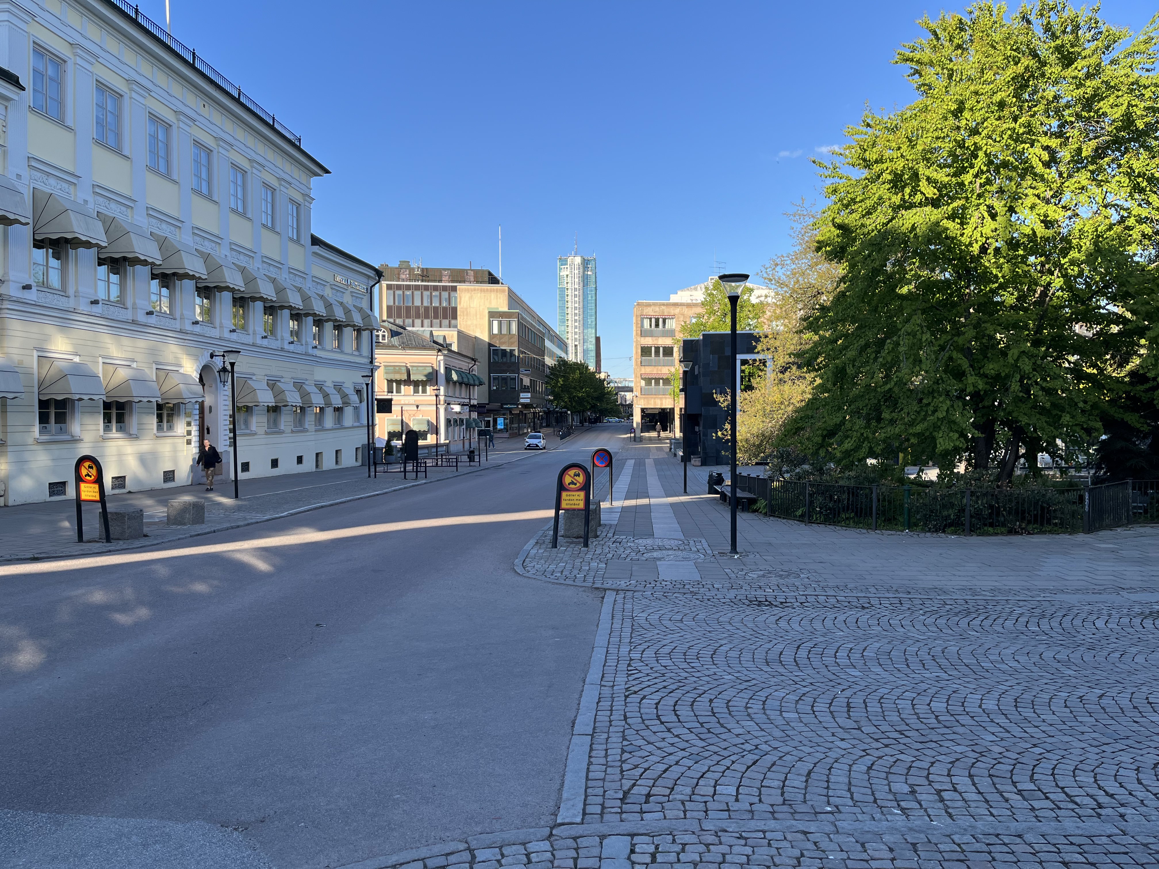 En gata i stadsmiljö med byggnader, träd och skyltar under en klarblå himmel. Till vänster syns en ljus byggnad med flera fönster och markiser. Längre fram på gatan står en hög byggnad och en person går på trottoaren till vänster.