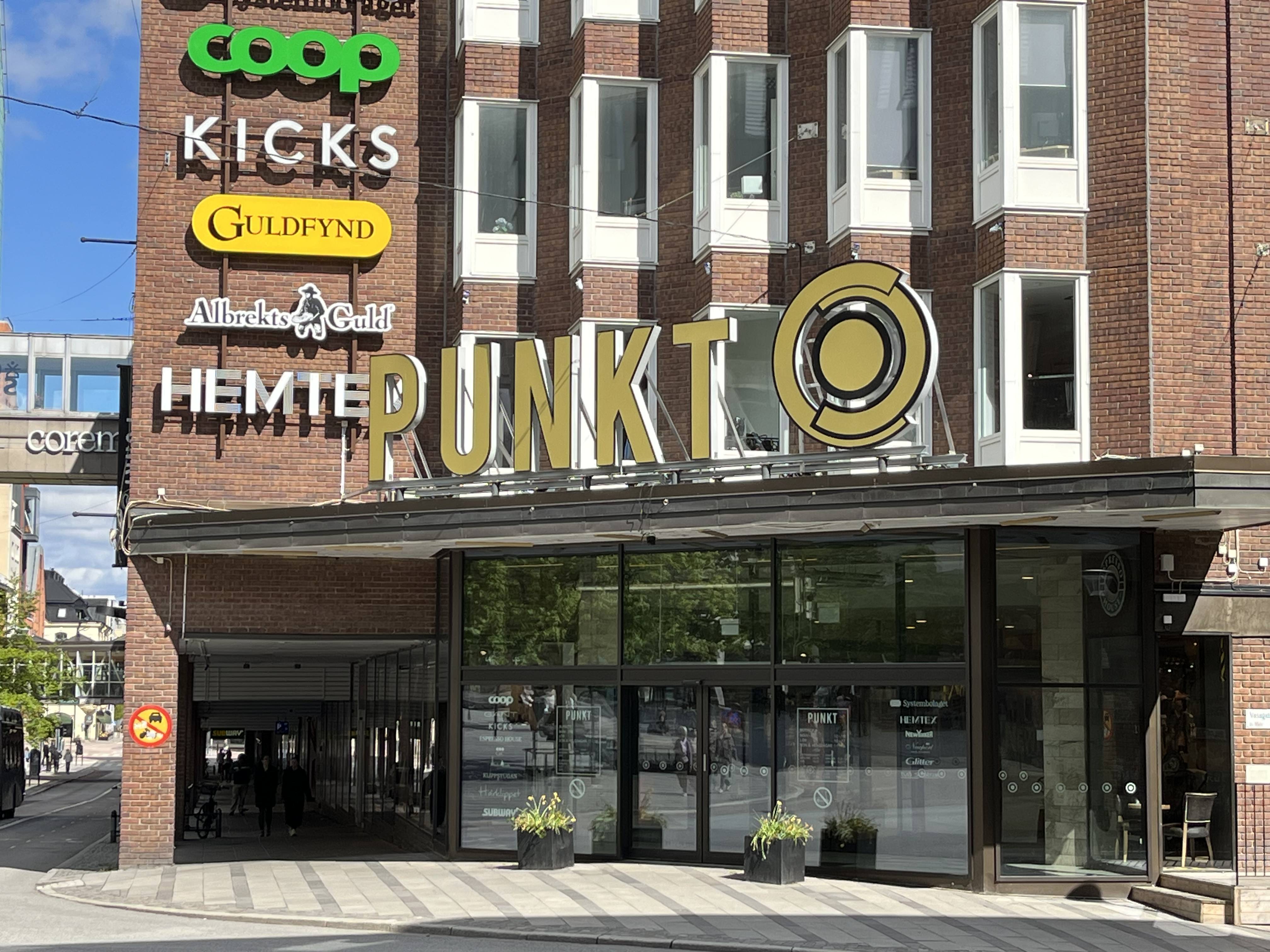 En fasad till ett tegelhus med flera butiksskyltar. Skyltar för Coop, Kicks, Guldfynd, Albrekts Guld, Hemtex och Punkt syns på byggnaden. Utanför entrén finns krukor med växter och det reflekteras grönska i fönstren. Ett bussfönster syns till vänster