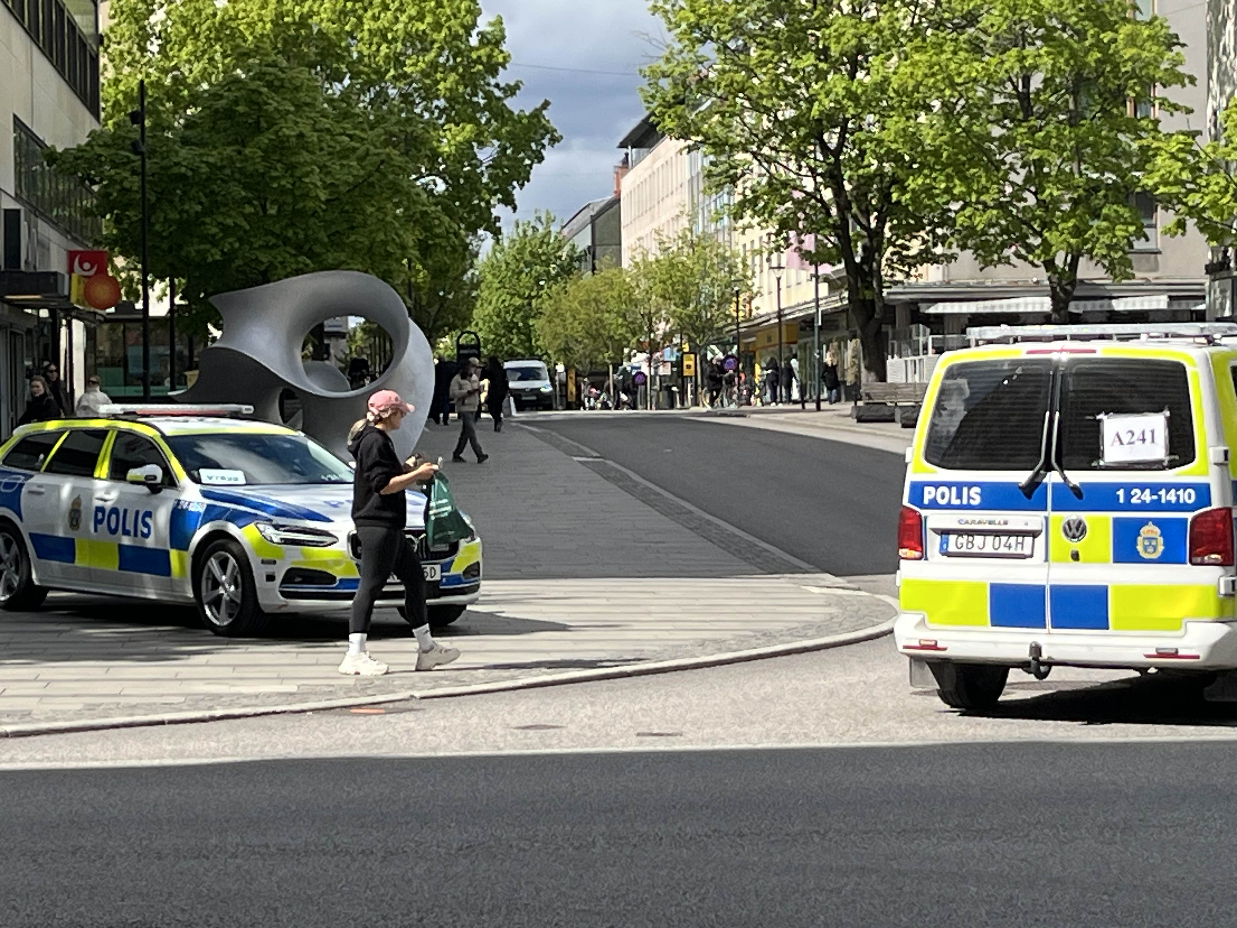 En gata i ett tätbebyggt område med flera personer och två parkerade polisbilar. Till vänster står en Volvo i polismålning och en person går i förgrunden med en påse. Till höger syns bakdelen av en VW skåpbil i polismålning. Gröna träd och byggnader