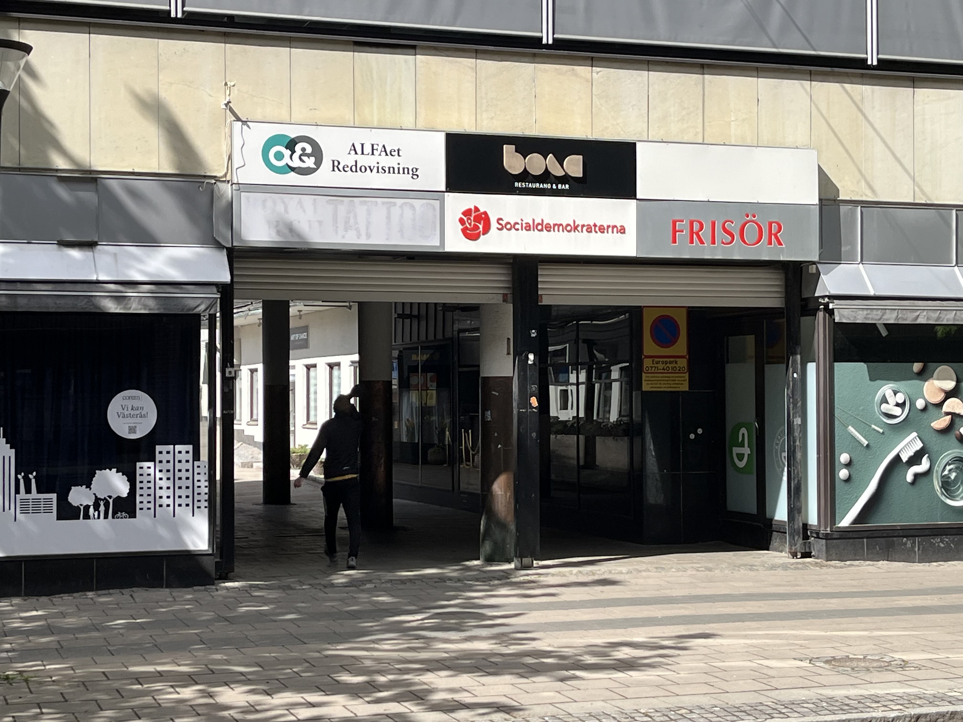 En person går under en portal med flera butiksskyltar. Skyltarna visar "ALFAet Redovisning", "bora Restaurang & Bar", "Socialdemokraterna avdelning Västerås", och "FRISÖR". Till vänster finns ett skyltfönster med texten "corem Vi kan Västerås!".