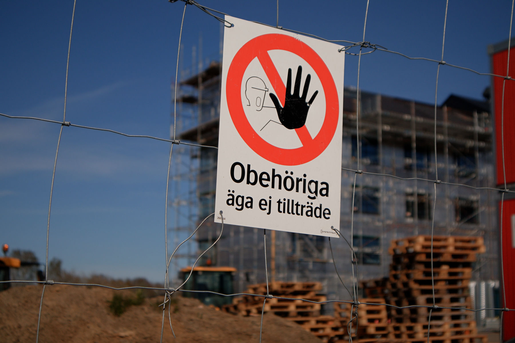 En bild som visar en skylt fäst vid ett stålstängsel. Skylten har en röd cirkel med ett svart streck diagonalt över en figur som signalerar stopp med handen som texten "Obehöriga äga ej tillträde". I bakgrunden syns en byggarbetsplats med ett oklart