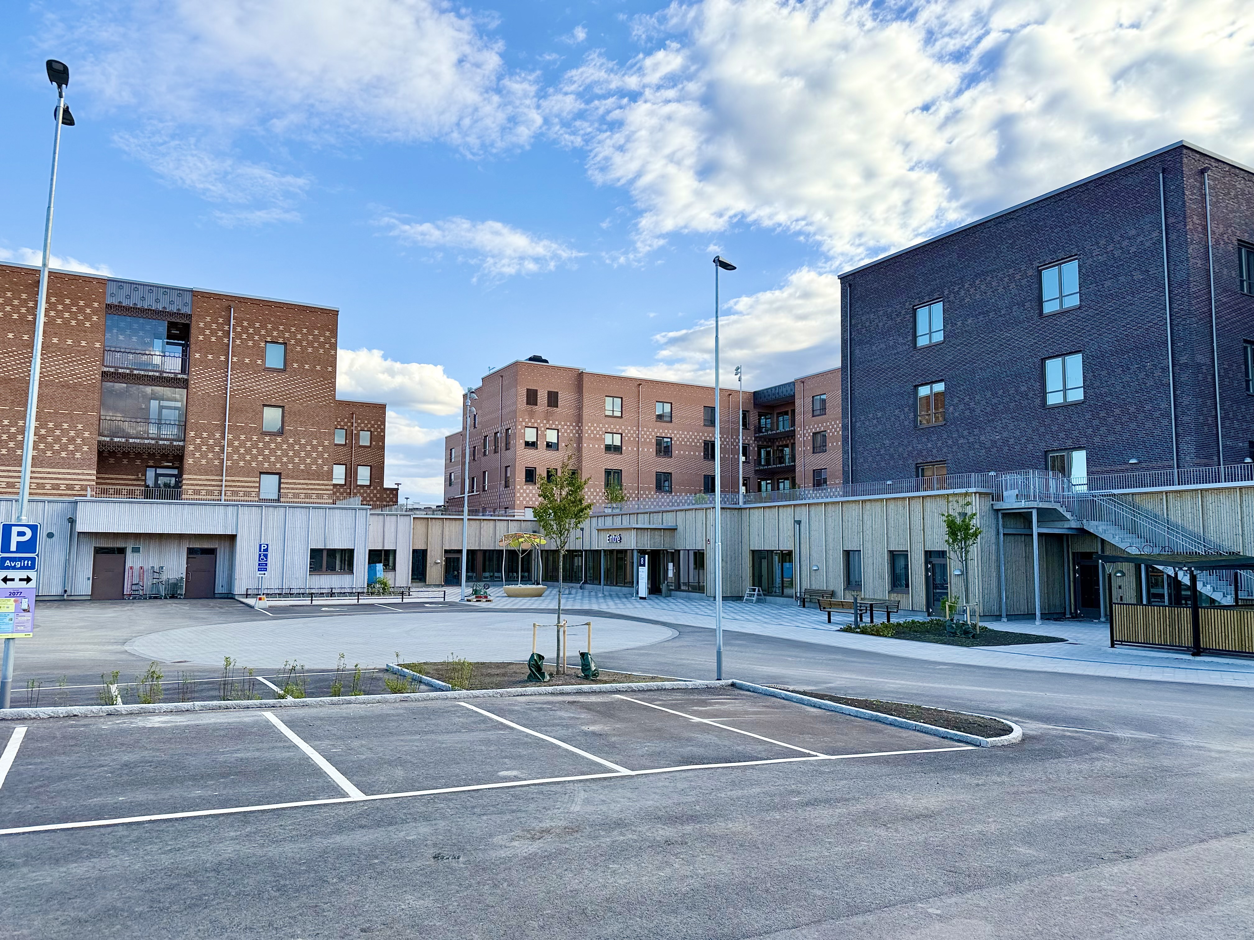 En bild som visar en modern byggnadskomplex med tegel- och träfasader under en blå himmel med vita moln. Framför byggnaderna finns en asfalterad parkeringsplats med vita markeringar och en gångväg. Mellan byggnaderna syns planteringar och en entré so