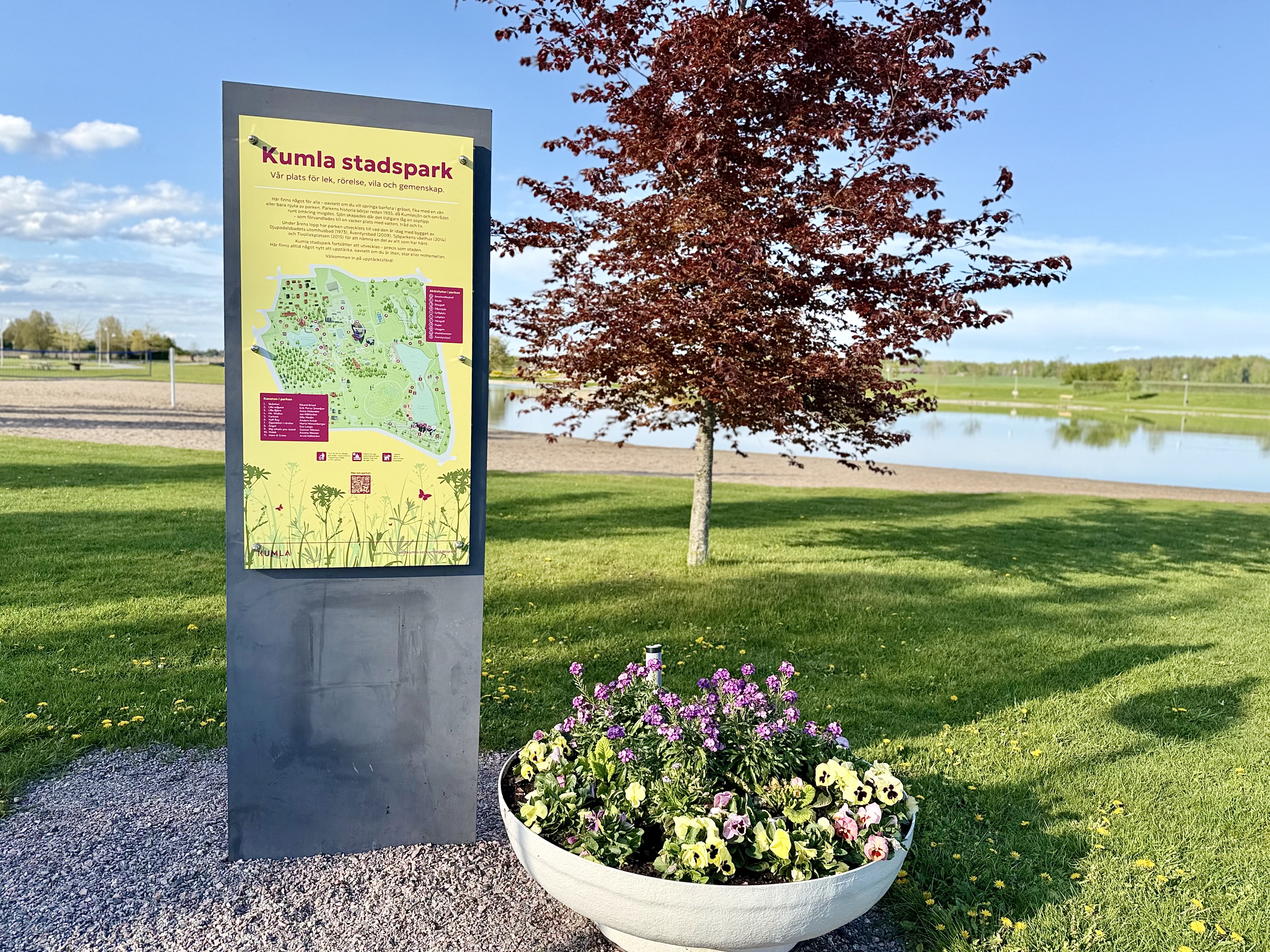 En vägkarta för Kumla stadspark avbildas nära ett landskap med blommor, träd och en sjö under en klarblå himmel. Kartan visar parkens layout och informationstext.