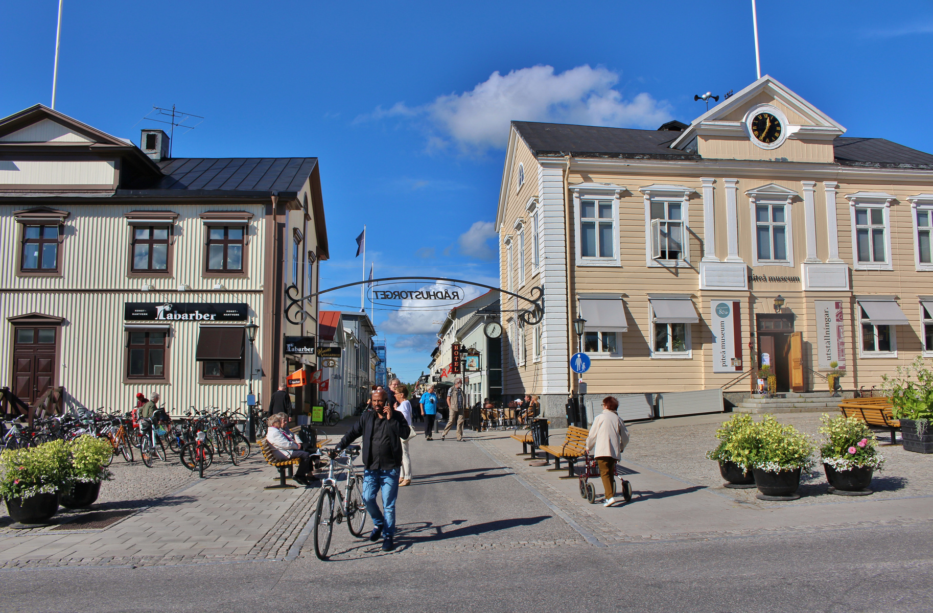 En gatuscen från en stadsmiljö med byggnader på båda sidor av en gata. Personer går och cyklar på gatan. En båge med texten "RÅDHUSTORGET" (spegelvänt) spänner över gatan. Till höger syns en ljusgul byggnad med en stor klocka på fasaden och texten "p