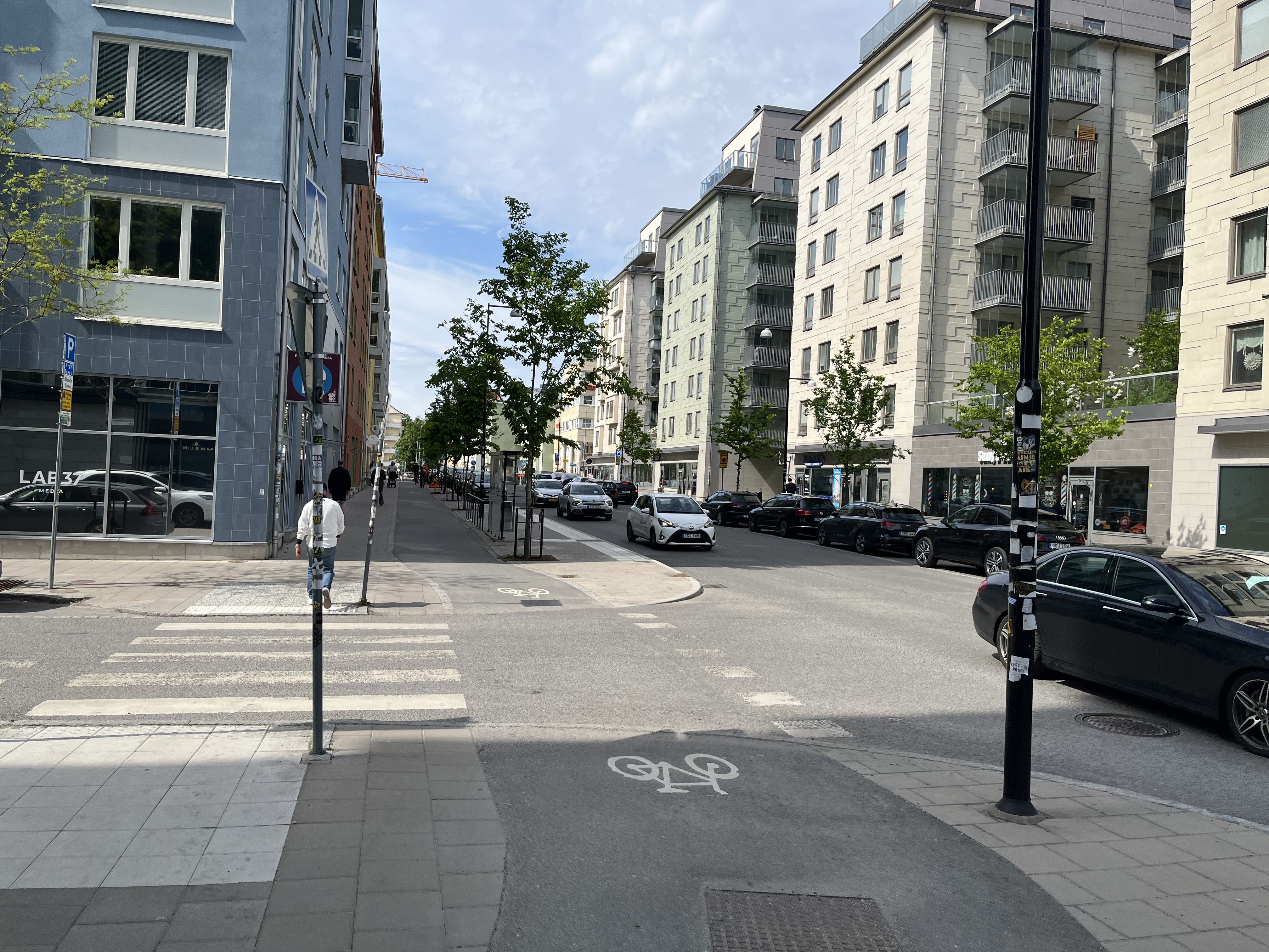 En gata i stadsmiljö med flerbostadshus på båda sidor. På gatan syns bilar parkerade längs med trottoarerna och i körfältet. Lövträden har full grönska, och himlen är molnig. En person går på trottoaren till vänster, bort från kameran.