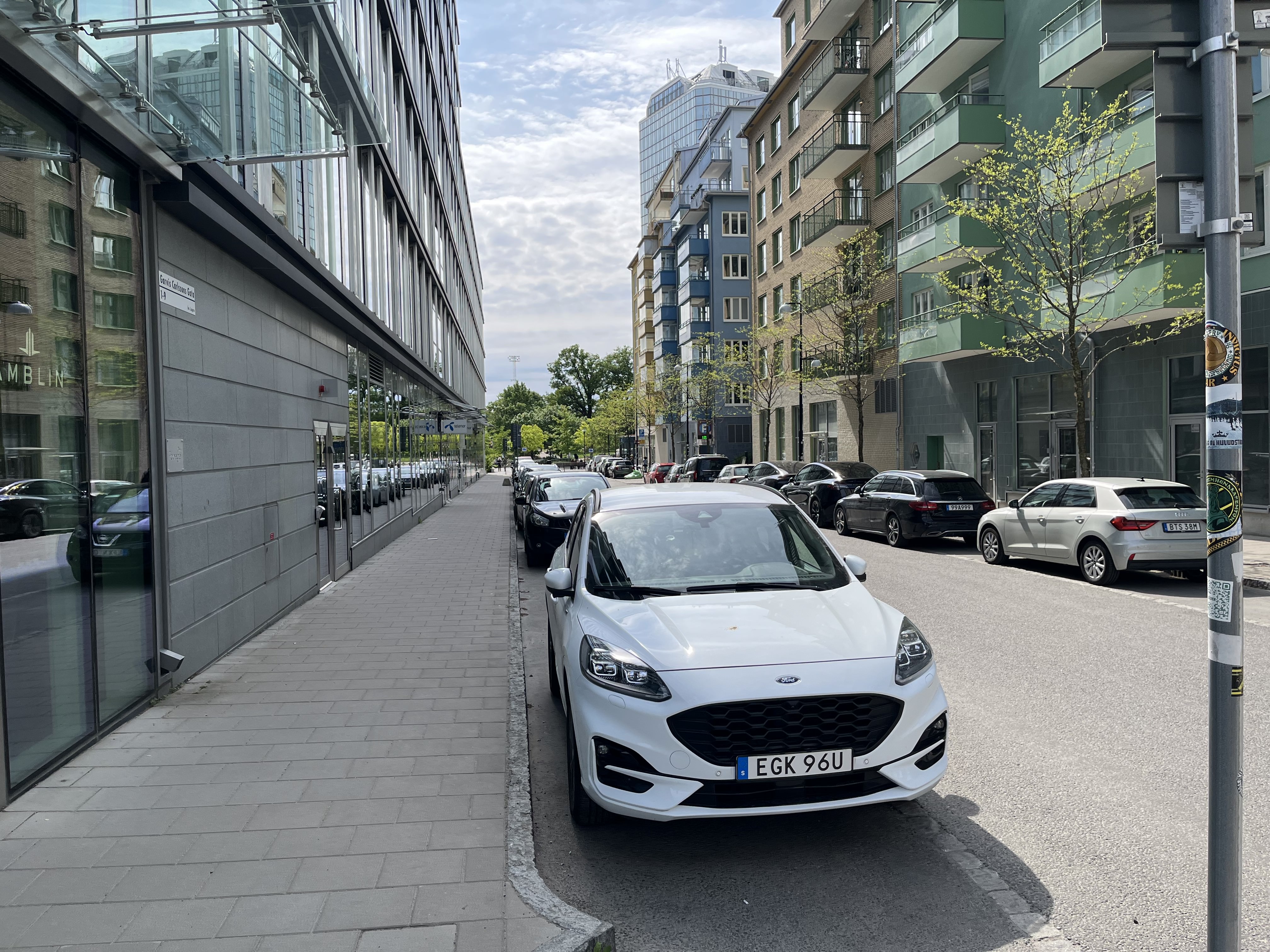 En gata inramad av byggnader med en rad parkerade bilar vid sidan. En vit personbil står i förgrunden till höger. På vänster sida av gatan syns en trottoar med en byggnad som har stora reflekterande fönster.