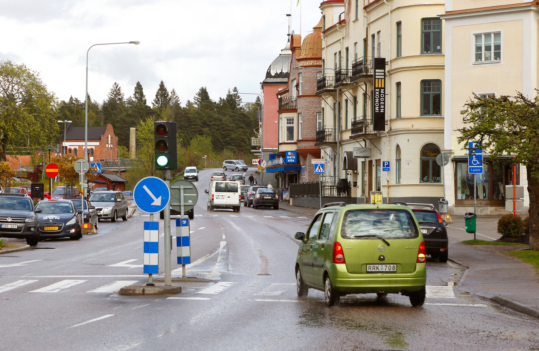 En gata i en stad med byggnader och fordon under en molnig dag. Ett gult hyreshus med balkonger syns till höger om bilden. En grön trafiksignal lyser, och en grön bil kör bort från kameran. Det är flera fordon på gatan, parkerade eller i rörelse.