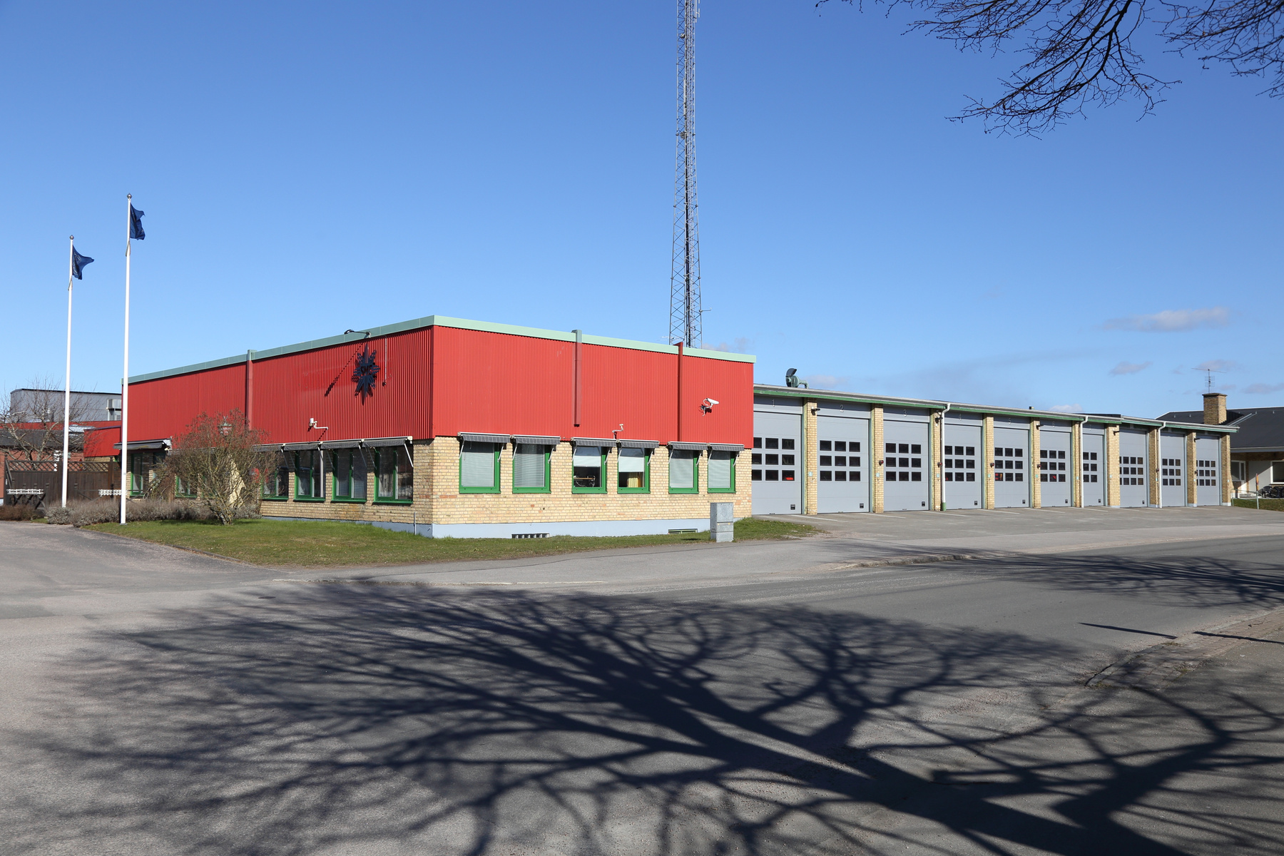 En röd och gul tegelbyggnad med gröna fönsterramar och flera garagedörrar står under en blå himmel. Framför byggnaden finns asfalt och en gräsyta med trädskuggor. Två flaggstänger med flaggor syns.