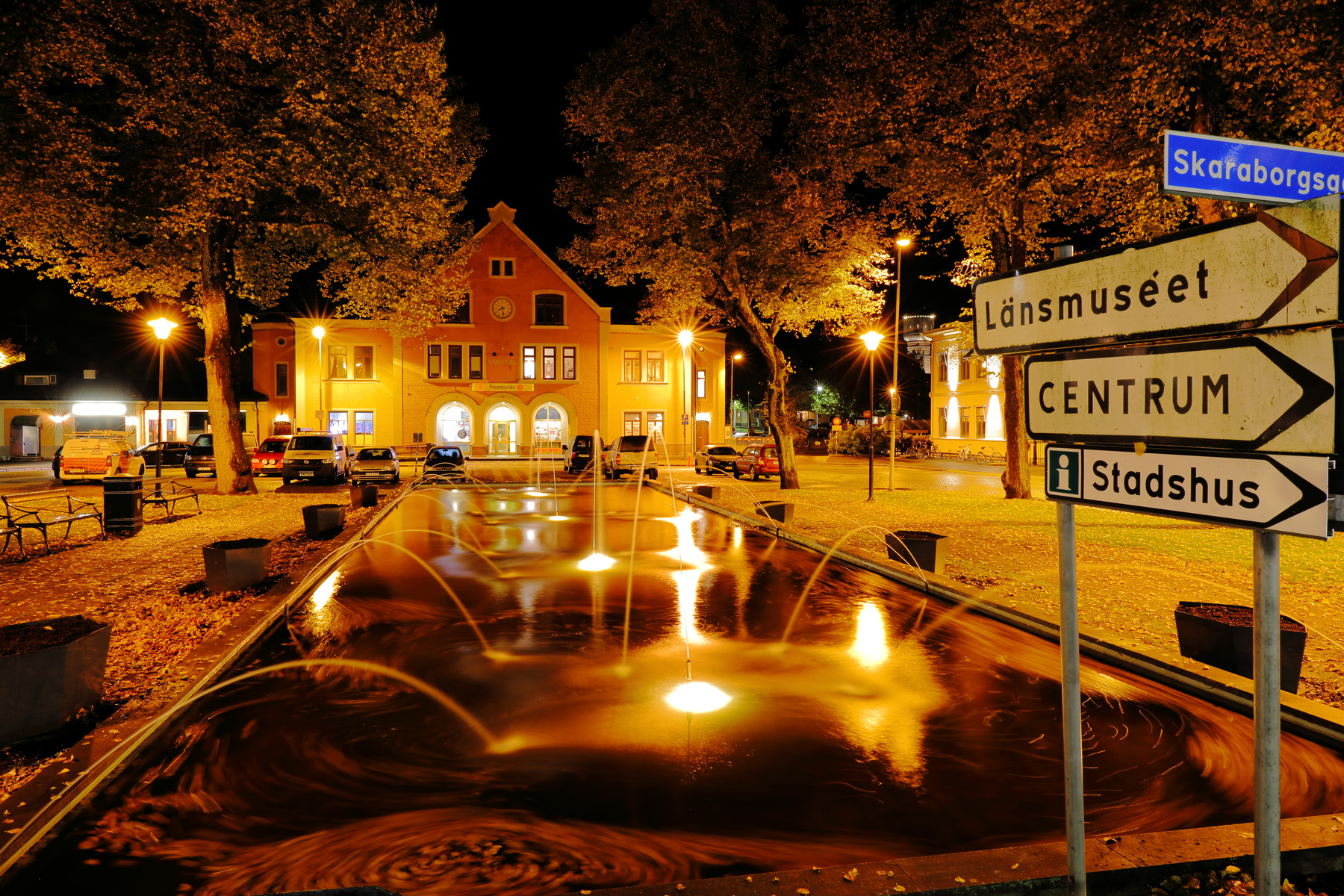 En nattbild visar en upplyst stadsmiljö med en stor byggnad i bakgrunden. Ett torg med lövfällande träd och en fontän med lysande vattenstrålar. Till höger syns en vägvisare med texten "Länsmuseet", "Centrum" och "Stadshus". Flera bilar är parkerade