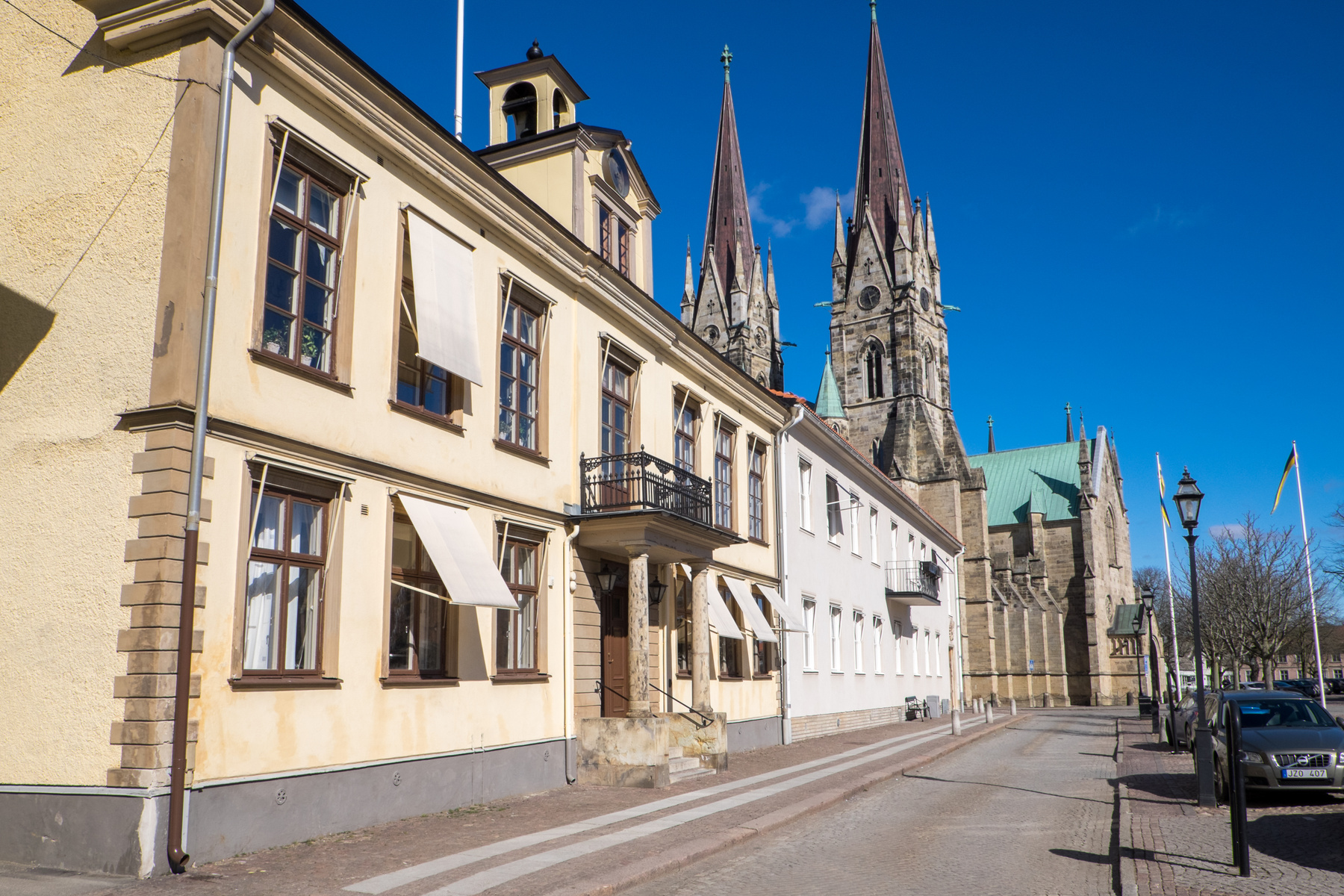 En gata med gatsten framför flera byggnader, inklusive en stor kyrka med två torn. Byggnaderna närmast kameran har fönster med ljusa markiser. På gatan står parkerade bilar och till höger syns nakna träd och en gatlykta. Himlen är klarblå och i förgr
