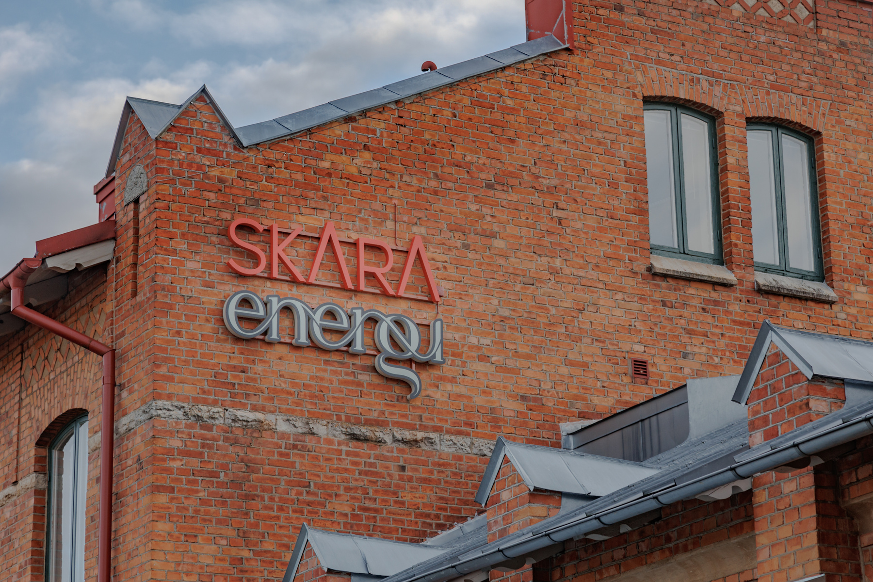 En stor del av en tegelbyggnad syns, med en skylt som lyder "SKARA energi" i rött och grått. Två välvda fönster med mörkgröna bågar är synliga, samt flera taklisterna och en röd stupränna. En blåsig himmel med moln ses i bakgrunden.