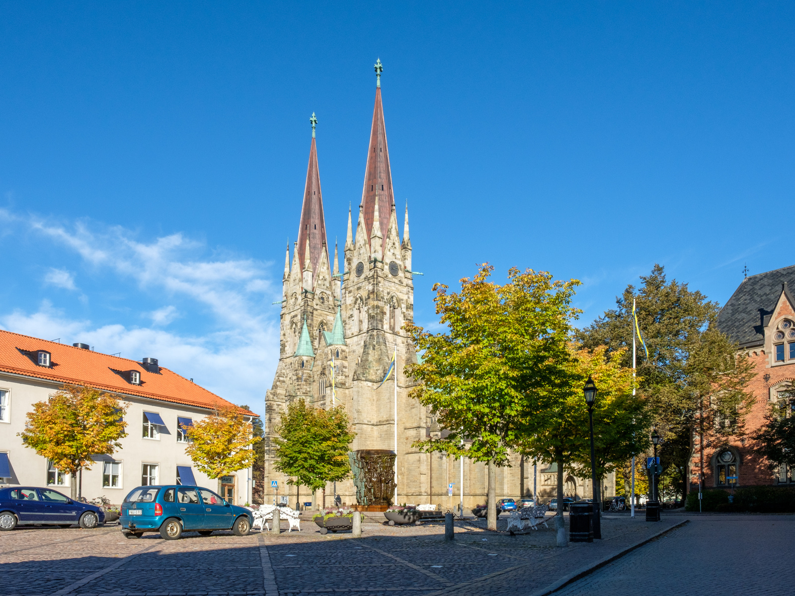 En stor stenkyrka med två höga, spetsiga torn är central i bilden. Framför kyrkan syns ett torg med gatsten, träd delvis med höstfärger, parkerade bilar och byggnader med röda tak. En flaggstång med en gul och blå vimpel syns till höger om kyrkan.