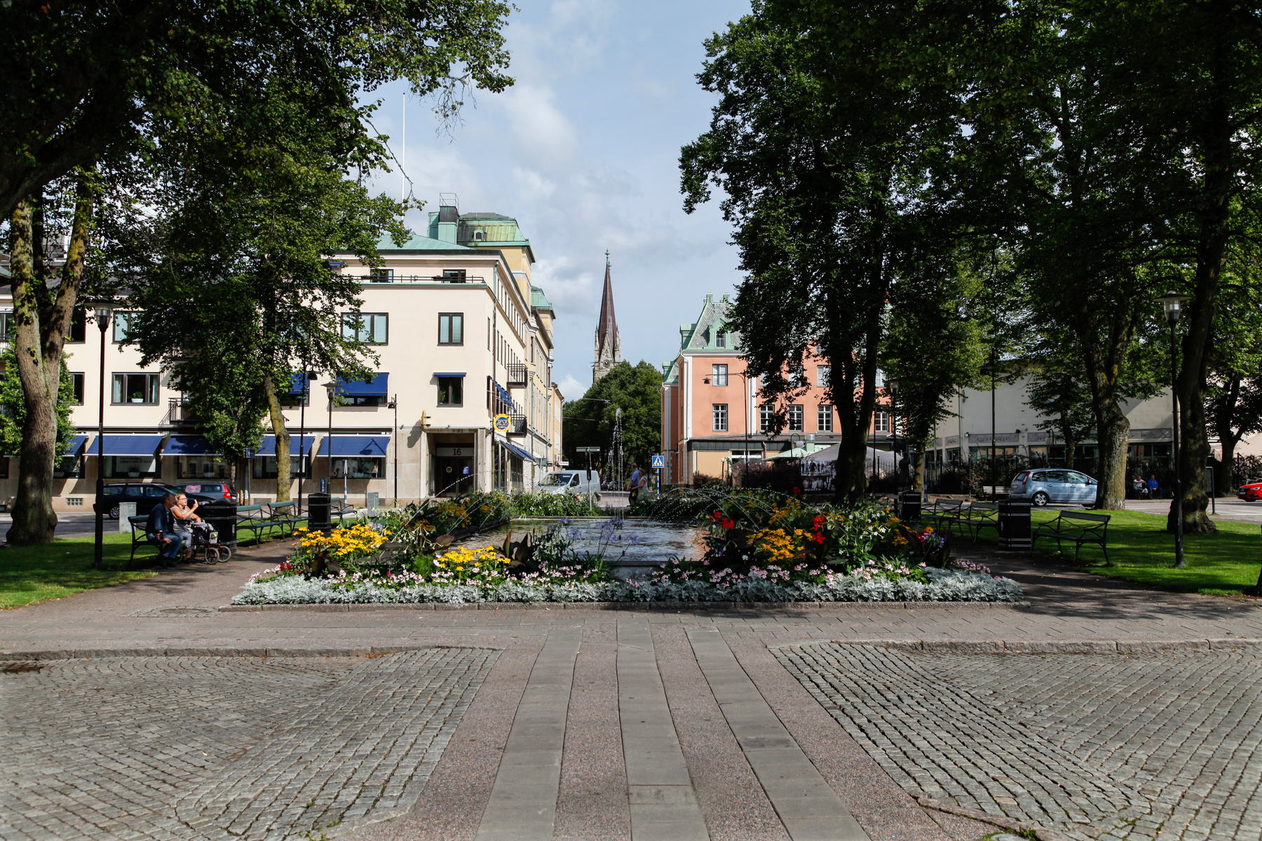 En central bild av en torgplats med en blommande fontän i förgrunden. Runt fontänen finns gräsmattor, träd och bänkar. På vardera sida syns byggnader, varav en större kyrkspira syns i bakgrunden. Två personer sitter på en bänk till vänster och en bil