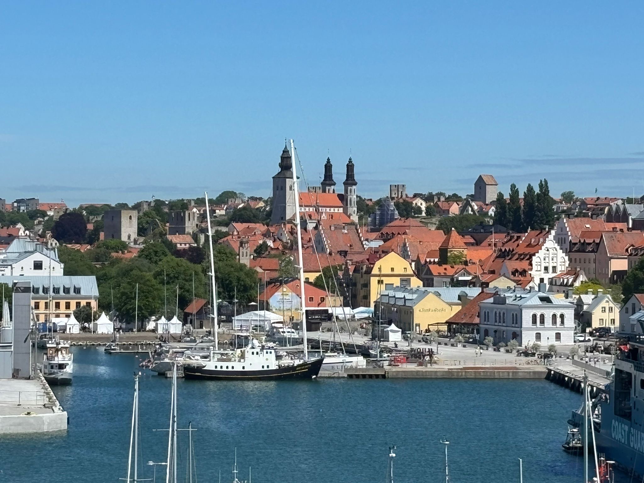 En översiktsbild av en hamn och en stad under en klarblå himmel. I förgrunden syns vatten med segelbåtar och förtöjda båtar. I bakgrunden reser sig stadens byggnader med röda tak, där bland annat kyrktorn och äldre ruiner är synliga. Längs kajen syns