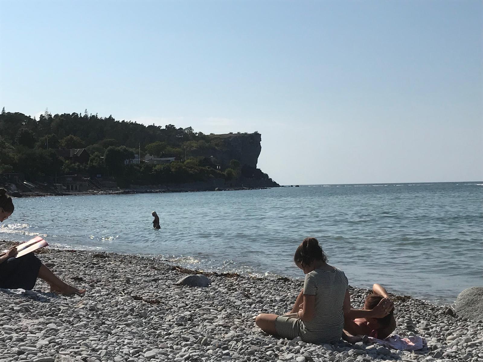 En bild tagen från en stenstrand med en bukt i förgrunden, omgiven av en brant, trädbevuxen kustlinje och en hög, kal klippformation i bakgrunden. Flera personer ses vid vattnet, varav en badar och andra sitter på stranden. Himmel är klarblå.