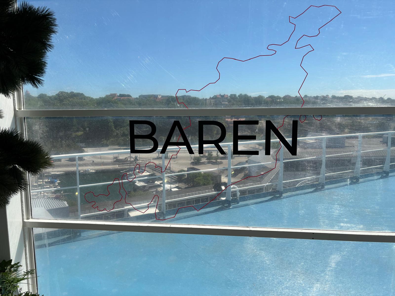 Bilden visar texten "BAREN" och en röd kontur av en landmassa, som troligen är Gotland, på en fönsterruta. Genom fönstret syns en marina eller mindre hamn, bebyggelse och grönska i bakgrunden under en klarblå himmel. Ett blått golv eller tak syns i f