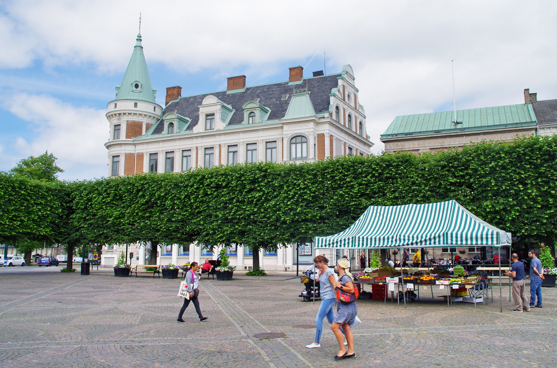 En utomhusbild som visar ett torg med flera människor. I förgrunden syns ytan med gatsten och i mitten en marknadsstånd med grön- och vitrandigt tak. Bakom ståndet finns träd med gröna bladverk och i bakgrunden en större byggnad med tinnar och torn.