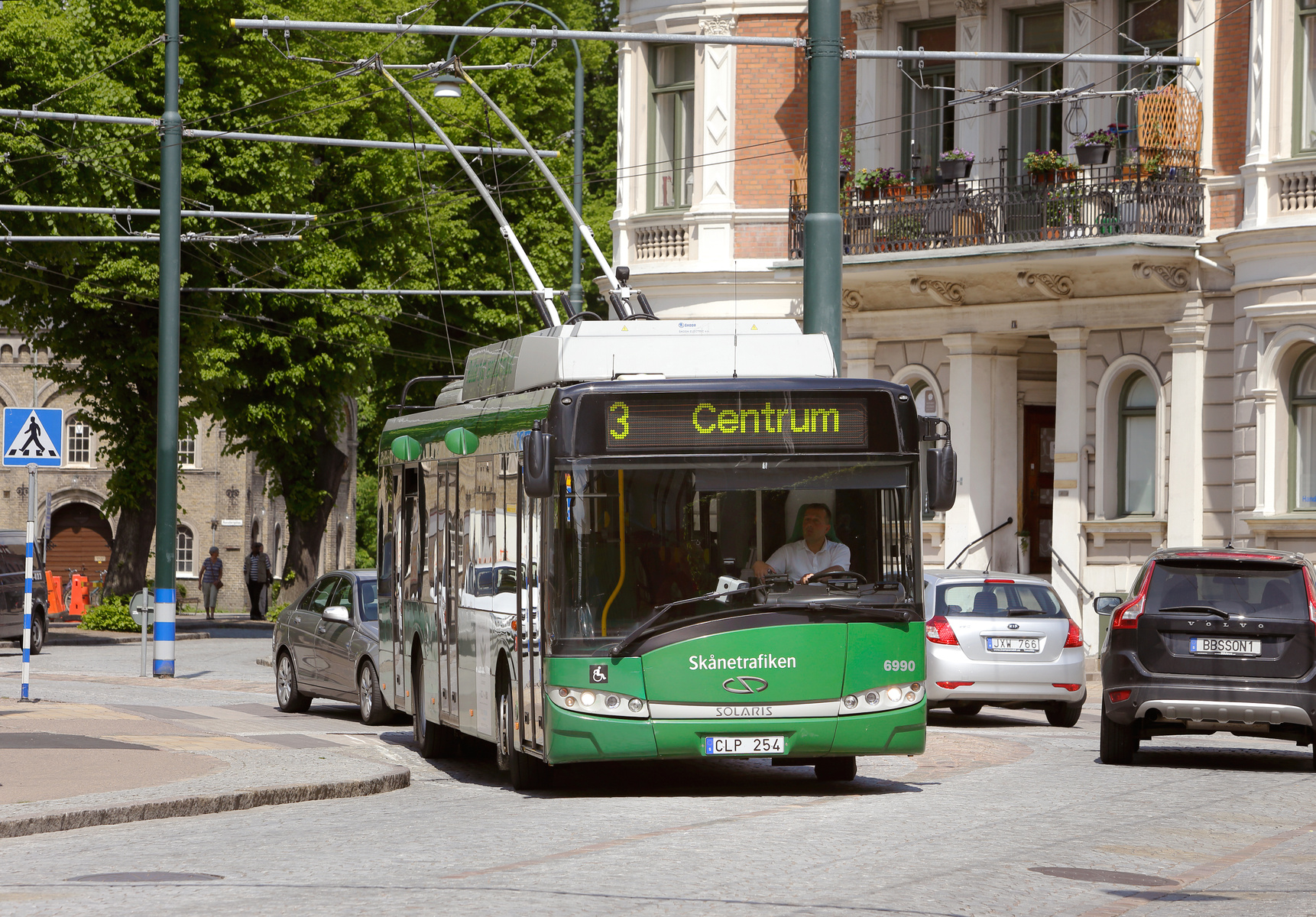 En grön buss märkt "Skånetrafiken" med destination "Centrum" kör på en kullerstensgata. I bakgrunden syns byggnader med utsmyckade balkonger och lummiga gröna träd. Förare och passagerare kan skymtas i bussen.