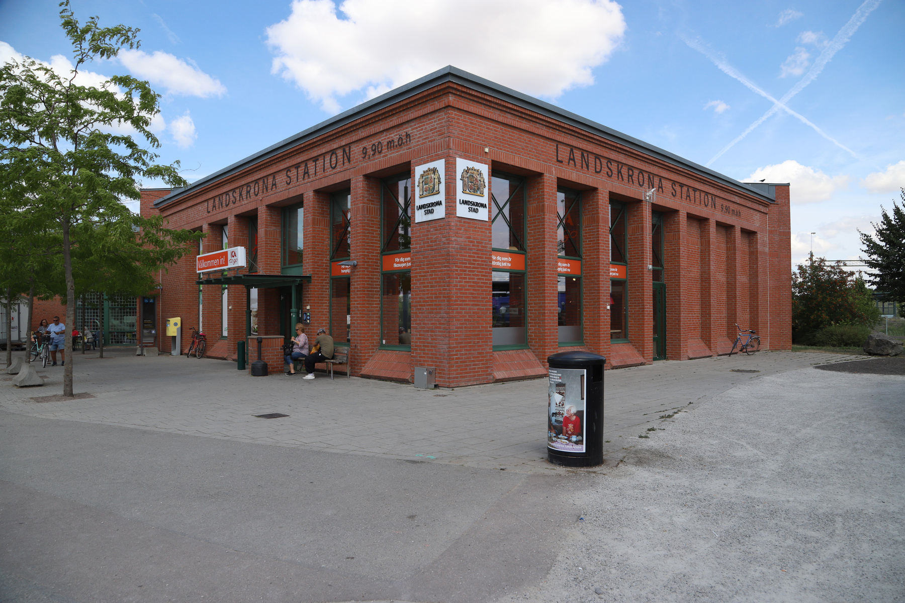 En röd tegelbyggnad med stora fönster och texten "LANDSKRONA STATION" på fasaden, under en blå himmel med vita moln och flygplansstråk. Flera personer syns runt byggnaden, en del sitter på en bänk. Träd och buskar syns till vänster och höger. På trot