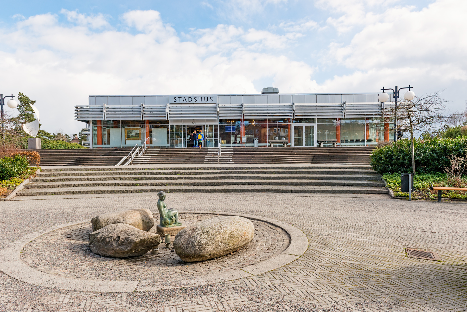 En modern byggnad med stora fönster och texten "STADSHUS" ovanför entrén. Framför byggnaden finns en bred trappa, och på marknivå en stenlagd yta med en skulptur föreställande en mänsklig figur bland stora stenar. Några personer syns i entrén.