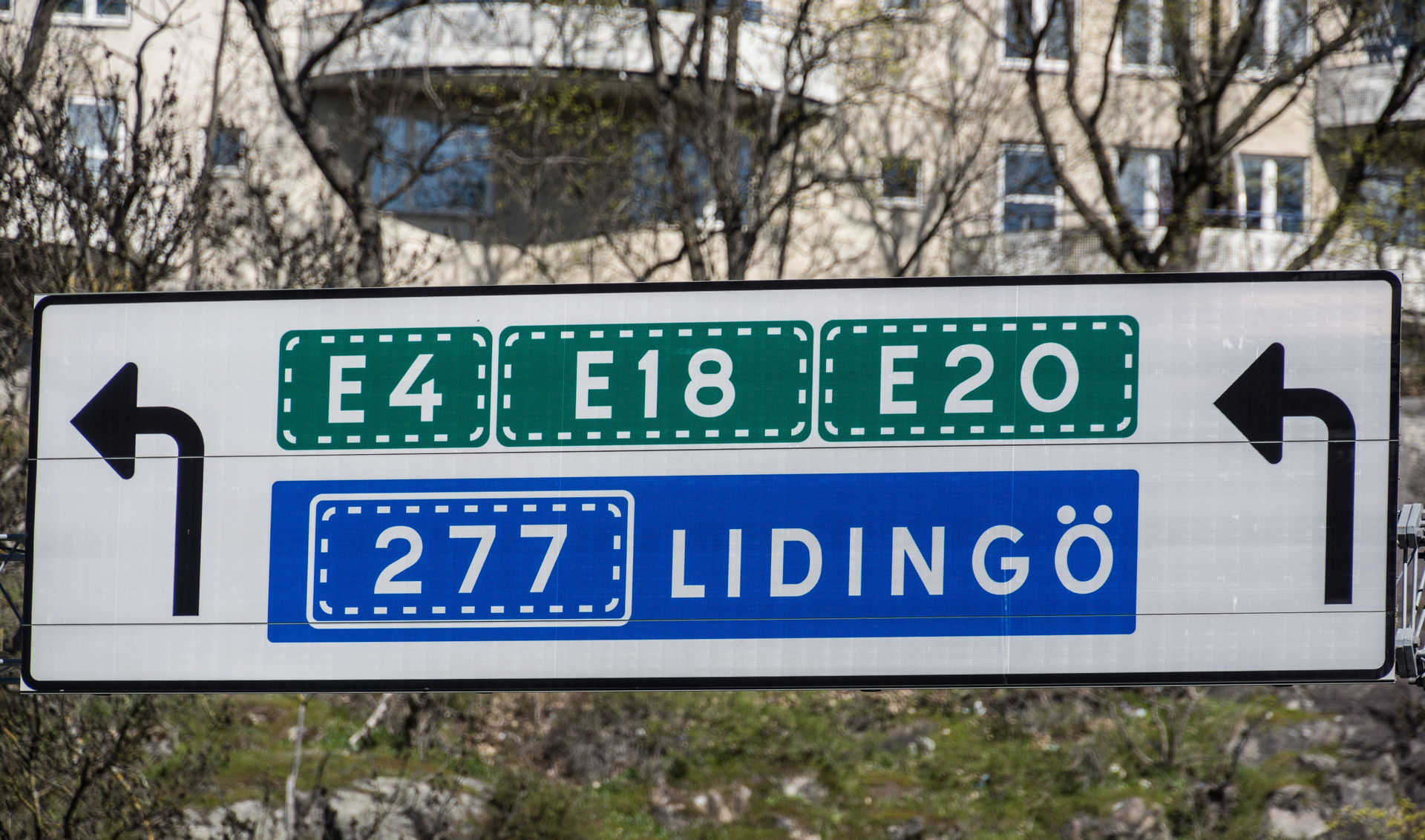 En vägskylt med information om vägar E4, E18, E20 och väg 277 mot Lidingö. Skylten har svarta pilsymboler som visar riktningen för vägarna. I bakgrunden syns delar av en byggnad och träd utan löv.
