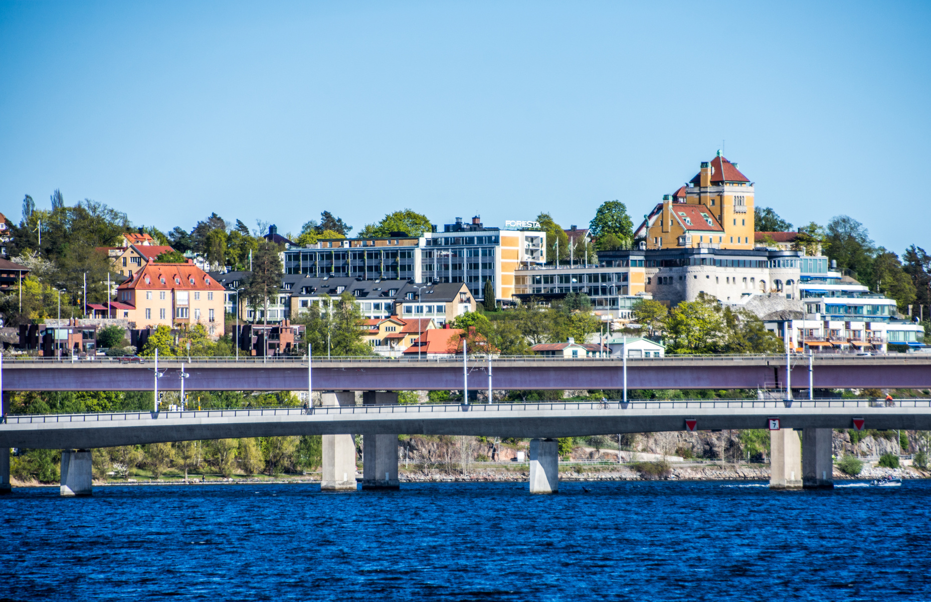 En bild tagen över vatten som visar en bebyggd kustlinje med broar i förgrunden och byggnader på en höjd i bakgrunden. En skylt med texten "FORESTA" är synlig.
