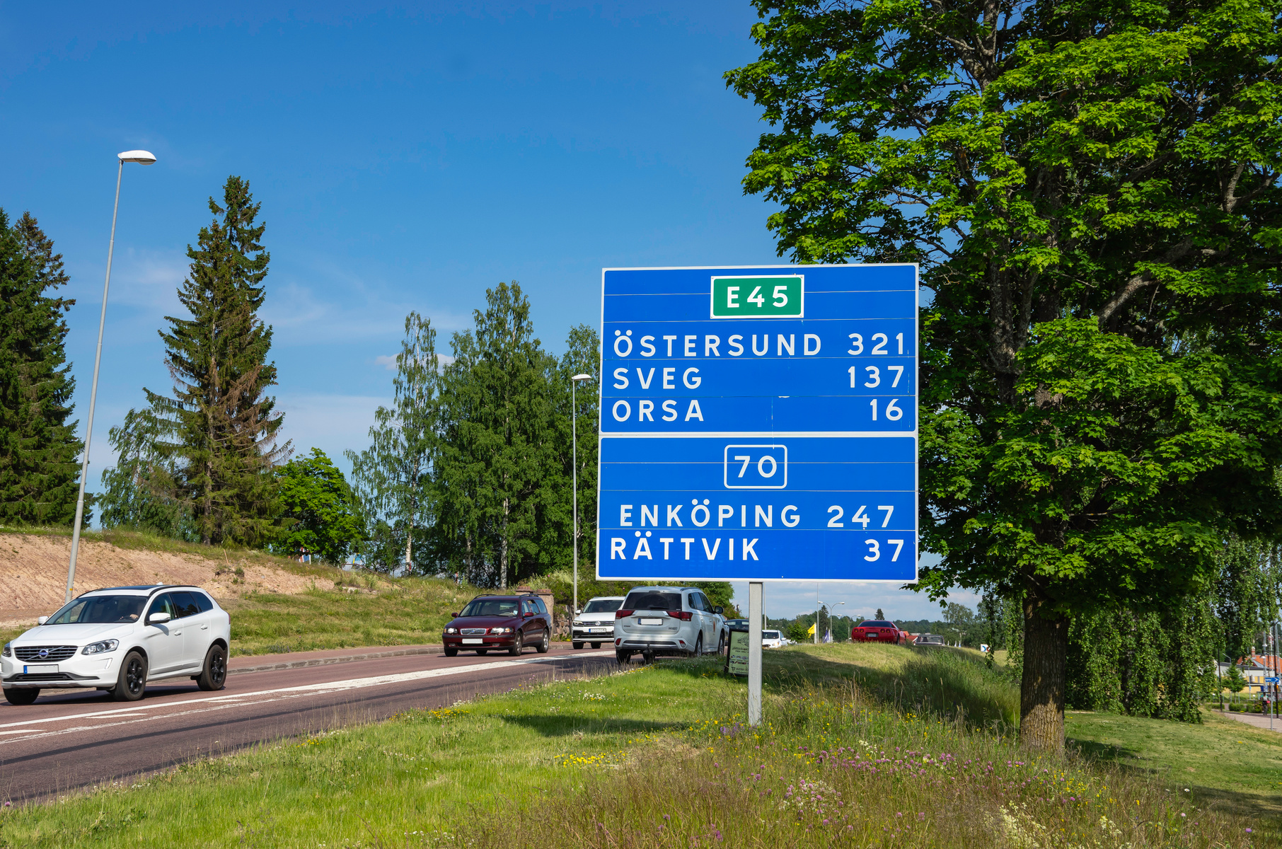 En blå vägskylt med vit text som visar riktning och avstånd till städerna Östersund, Sveg, Orsa, Enköping och Rättvik vid E45 och väg 70. Bilar färdas på en väg som kantas av gräs och träd under en blå himmel.