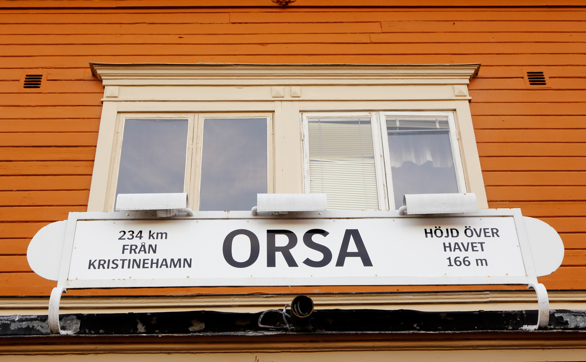 En vit skylt med svart text visar information om orten Orsa, inklusive avstånd (234 km från Kristinehamn) och höjd över havet (166 m). Skylten är monterad under ett fönsterparti med två fönster i en orange träfasad med liggande panel. Ett persienn sy