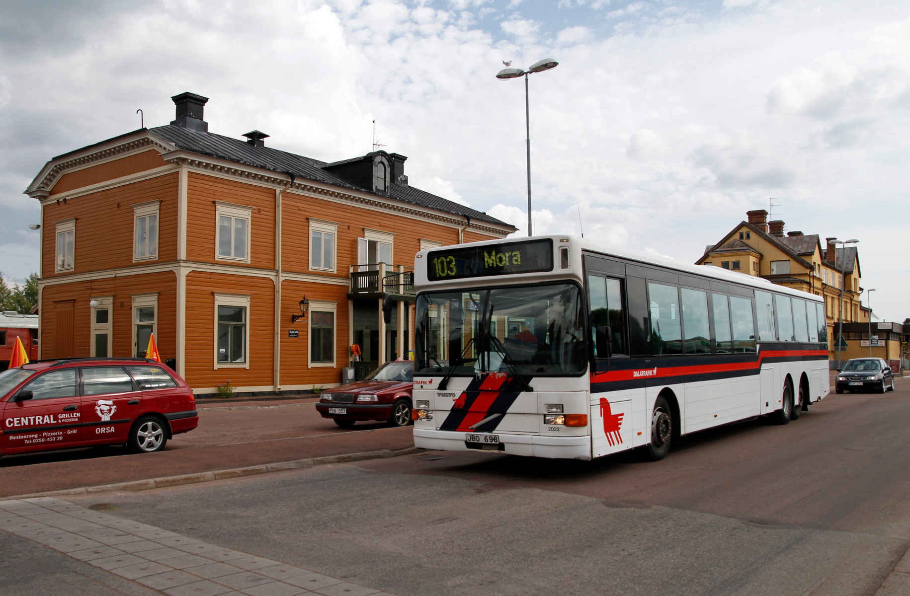 En vit ledbuss med röda och svarta detaljer svänger i en gatukorsning framför en ljusbrun byggnad med vita knutar och fönster. I bakgrunden syns fler byggnader och en molnig himmel. En röd kombibil med texten "CENTRAL GRILLEN" och en röd personbil sy